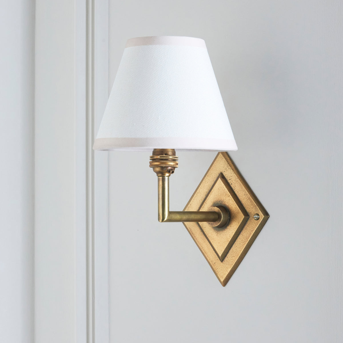 Endon Diamond Angle wall light unlit on a pale grey blue panelled wall, antique brass diamond backplate and crisp white Ivy shade