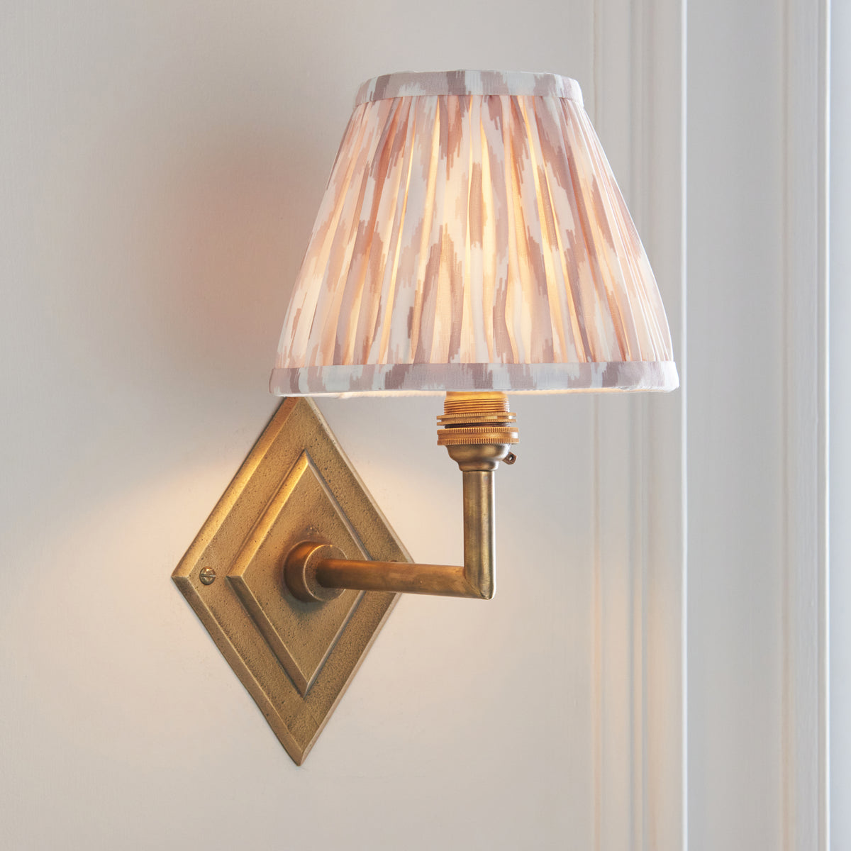 Lit Endon Diamond Angle wall light on cream panelling, close view of the brass diamond backplate, angled arm and pleated ikat shade