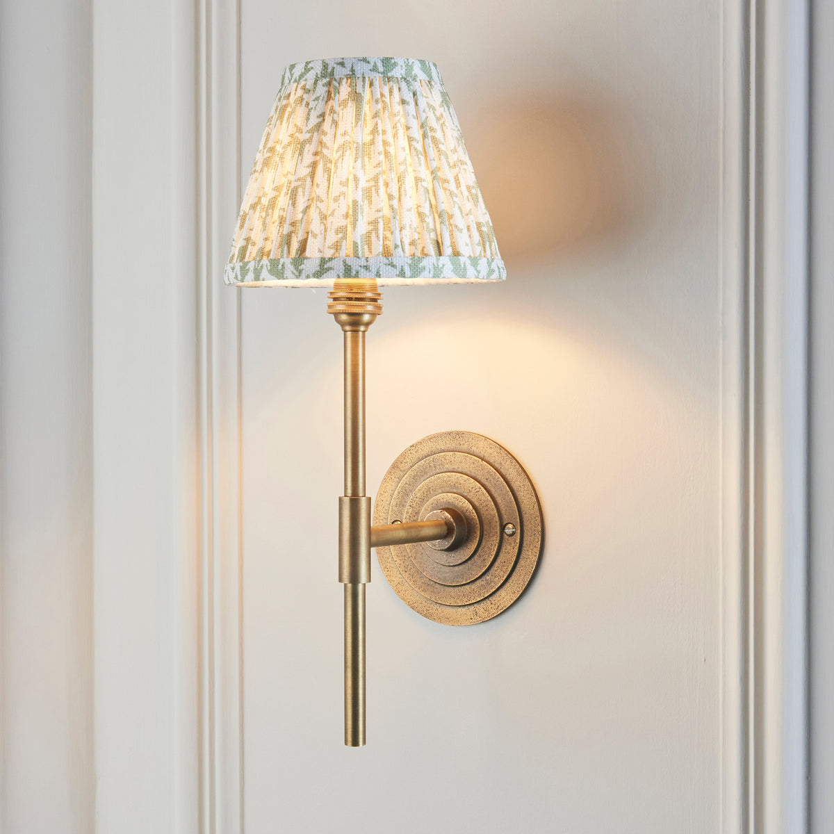 Endon Halo Tall antique brass wall light lit on a cream panelled wall, green leaf print pleated shade glowing warmly