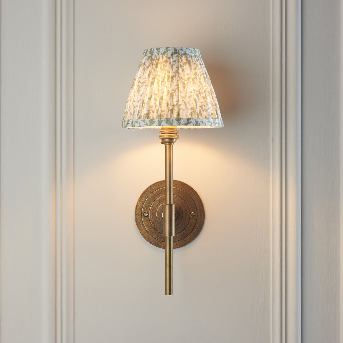 Endon Halo Tall wall light lit on a cream panelled wall, brass stem centred over the ringed backplate, green leaf shade