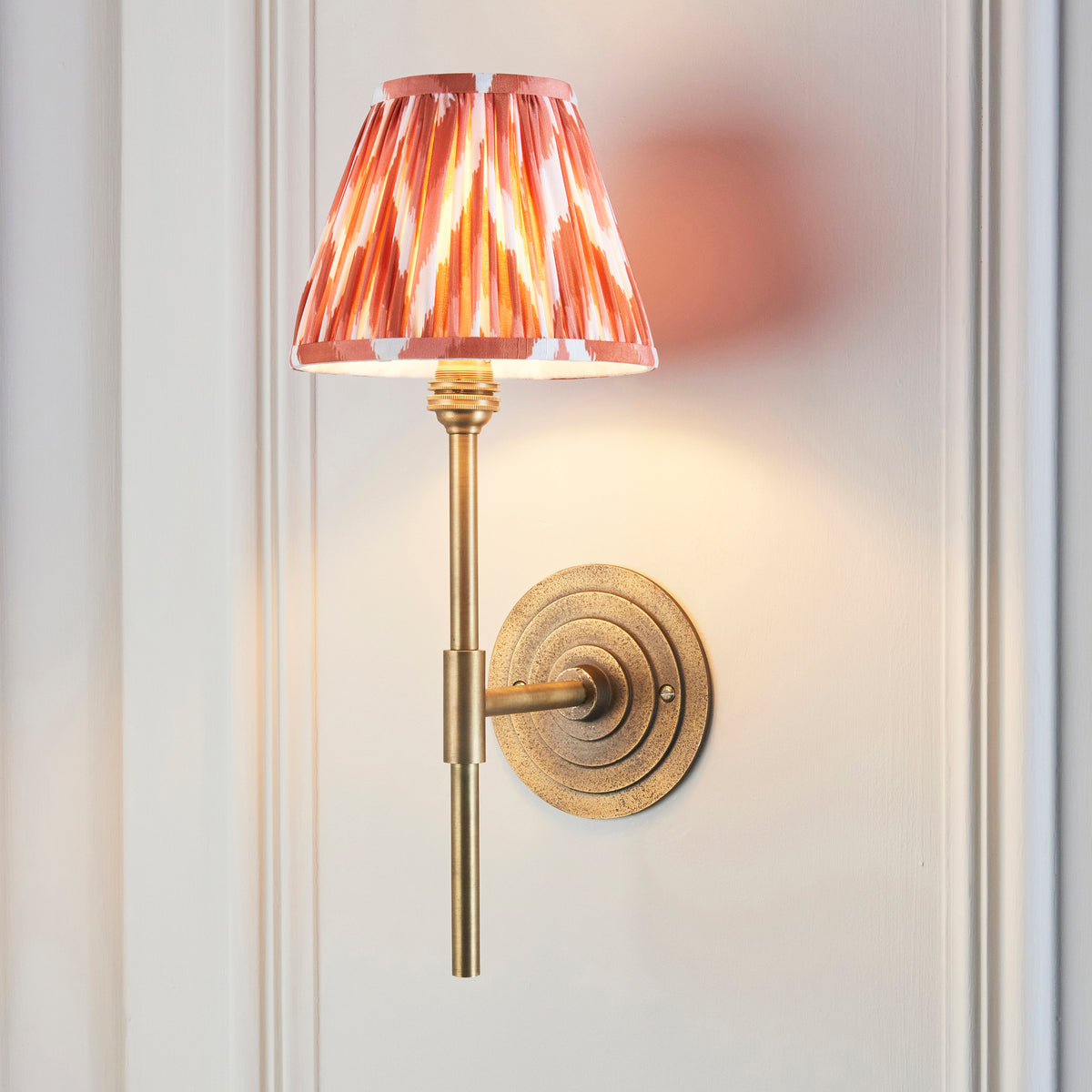 Endon Halo Tall antique brass wall light lit, apricot orange zigzag shade glowing warm on a cream panelled wall