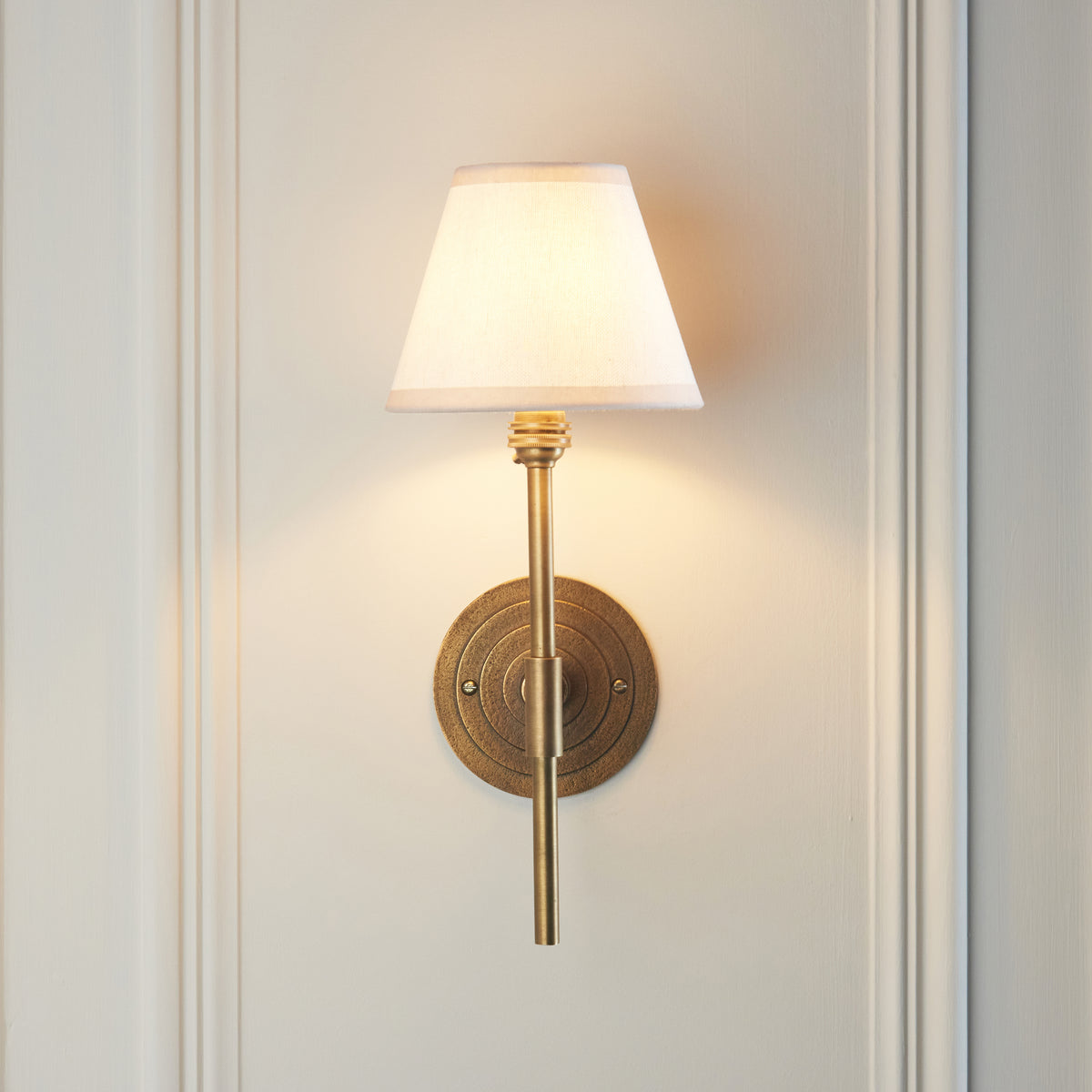 Endon Halo Tall wall light lit on a cream panelled wall, stepped antique brass backplate centred behind the straight brass column