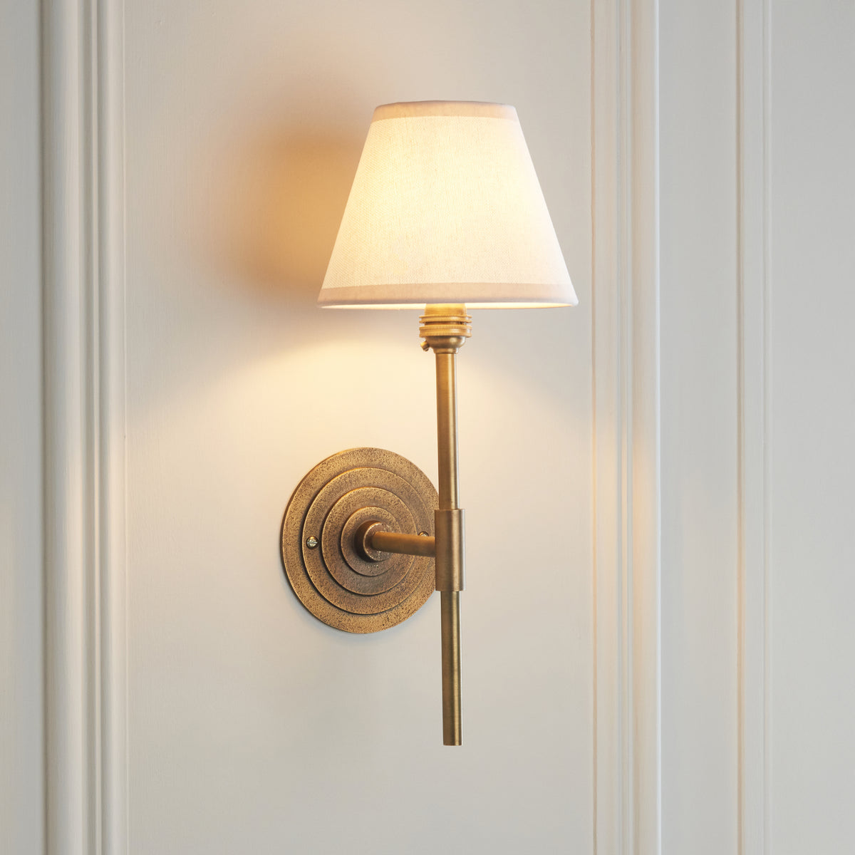 Endon Halo Tall antique brass wall light lit, casting a soft pool of light across a cream panelled wall around the Ivy shade