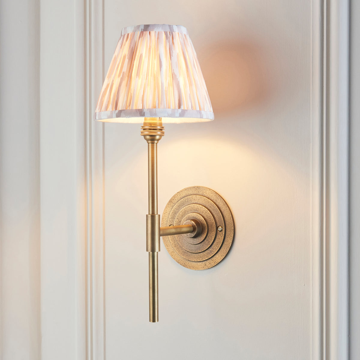 Endon Halo Tall antique brass wall light lit on a cream panelled wall, the neutral Ikat shade glowing above the ringed backplate