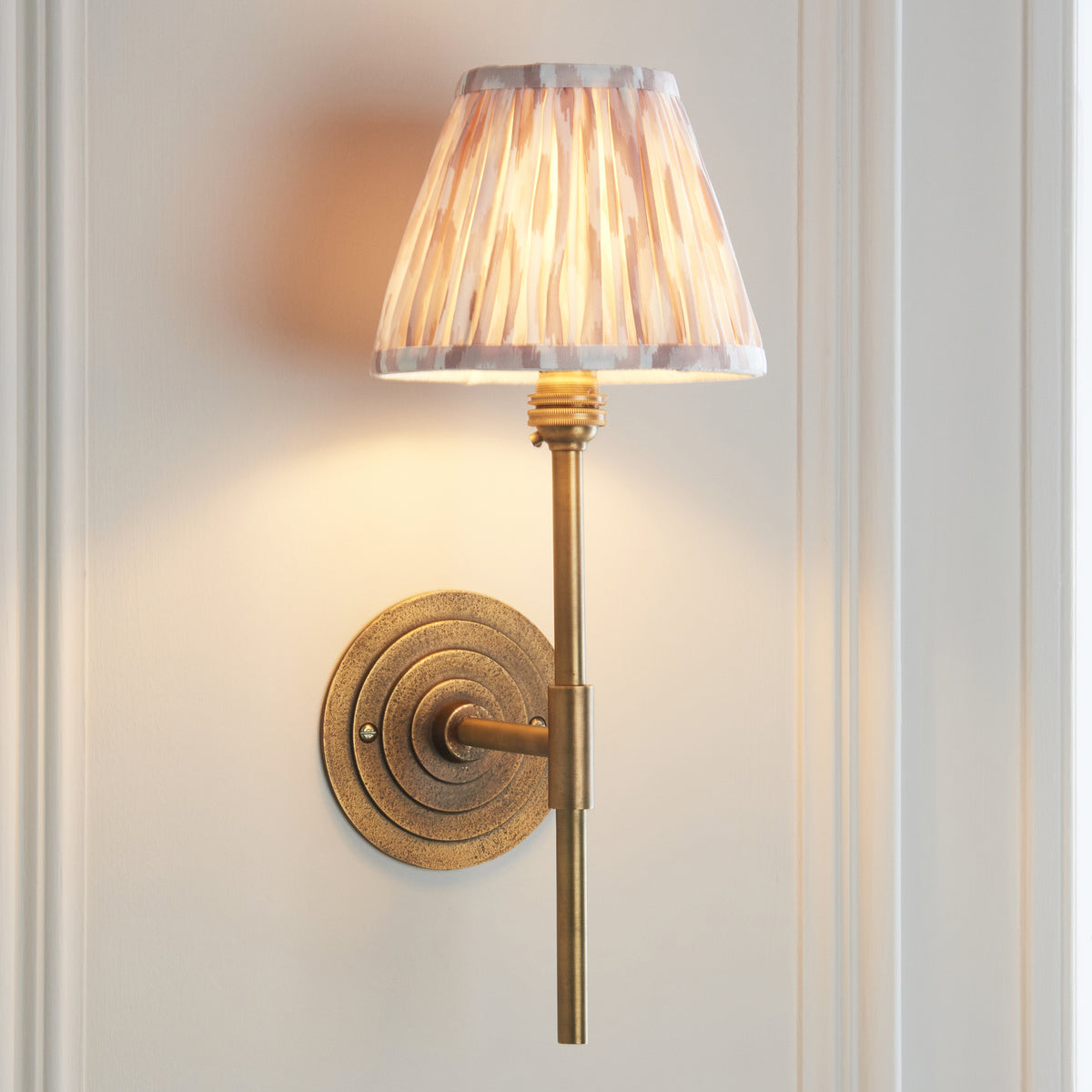 Endon Halo Tall wall light lit on a cream panelled wall, short brass arm reaching from the ringed backplate to the tall stem and pleated Ikat shade