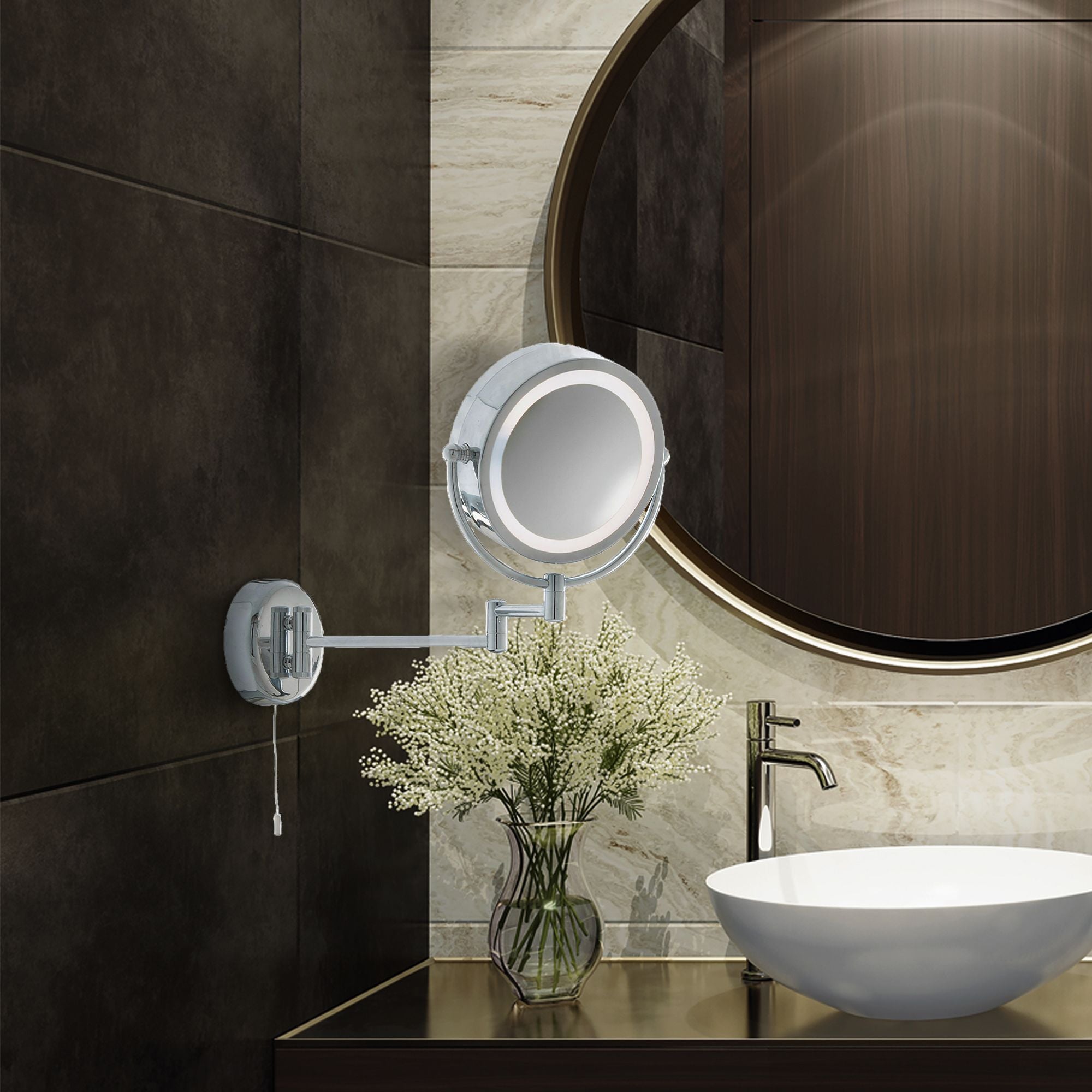 Searchlight illuminated chrome bathroom mirror mounted on a dark tiled wall beside a round mirror, basin and vase of flowers