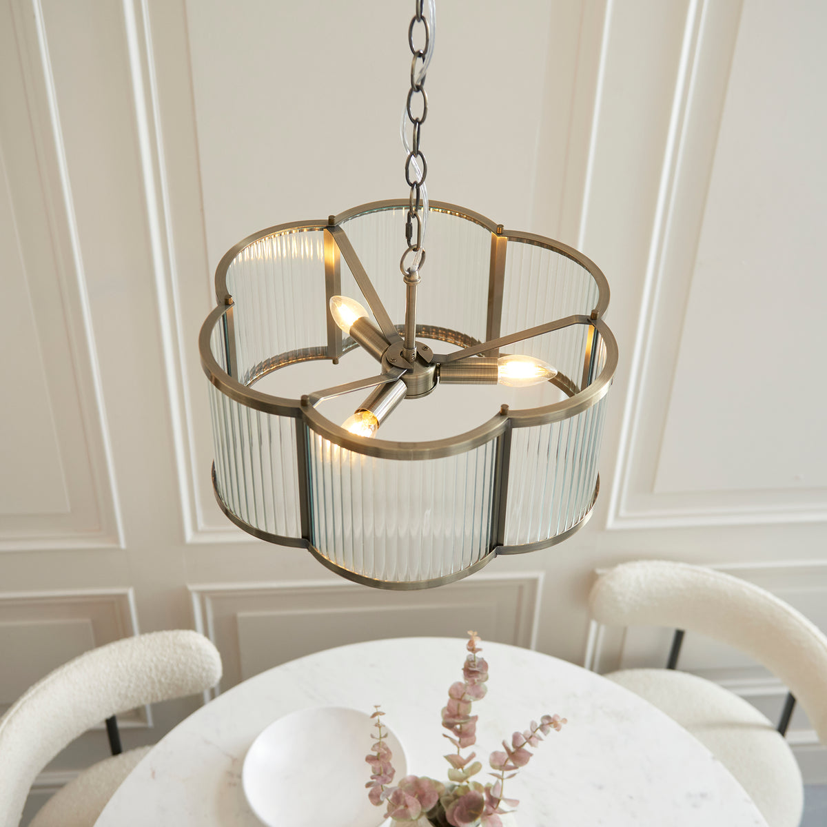 Angled view from below the lit Ridgeton Scallop pendant showing the candelabra bulbs inside the ribbed glass panels above a dining table set with eucalyptus stems and chairs.