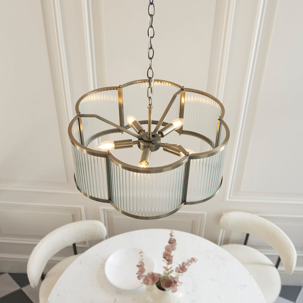 Angled lifestyle view of the lit Ridgeton Scallop five light pendant showing the brass spoke frame and candelabra bulbs inside the glass panels above a table with flowers.