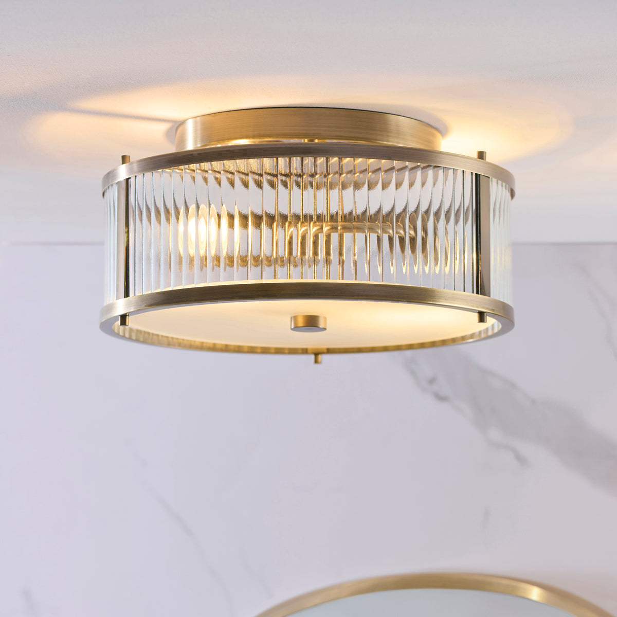 Close up lifestyle shot of the Endon Ridgeton IP44 flush ceiling light in antique brass with clear ribbed glass panels lit against a marble effect bathroom wall
