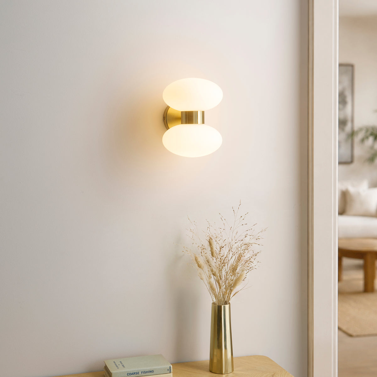 Lit lifestyle shot of the Endon Rossano 2 light wall light in warm brass with opal glass shades on a grey wall with a ceramic vase and stacked books on a shelf below