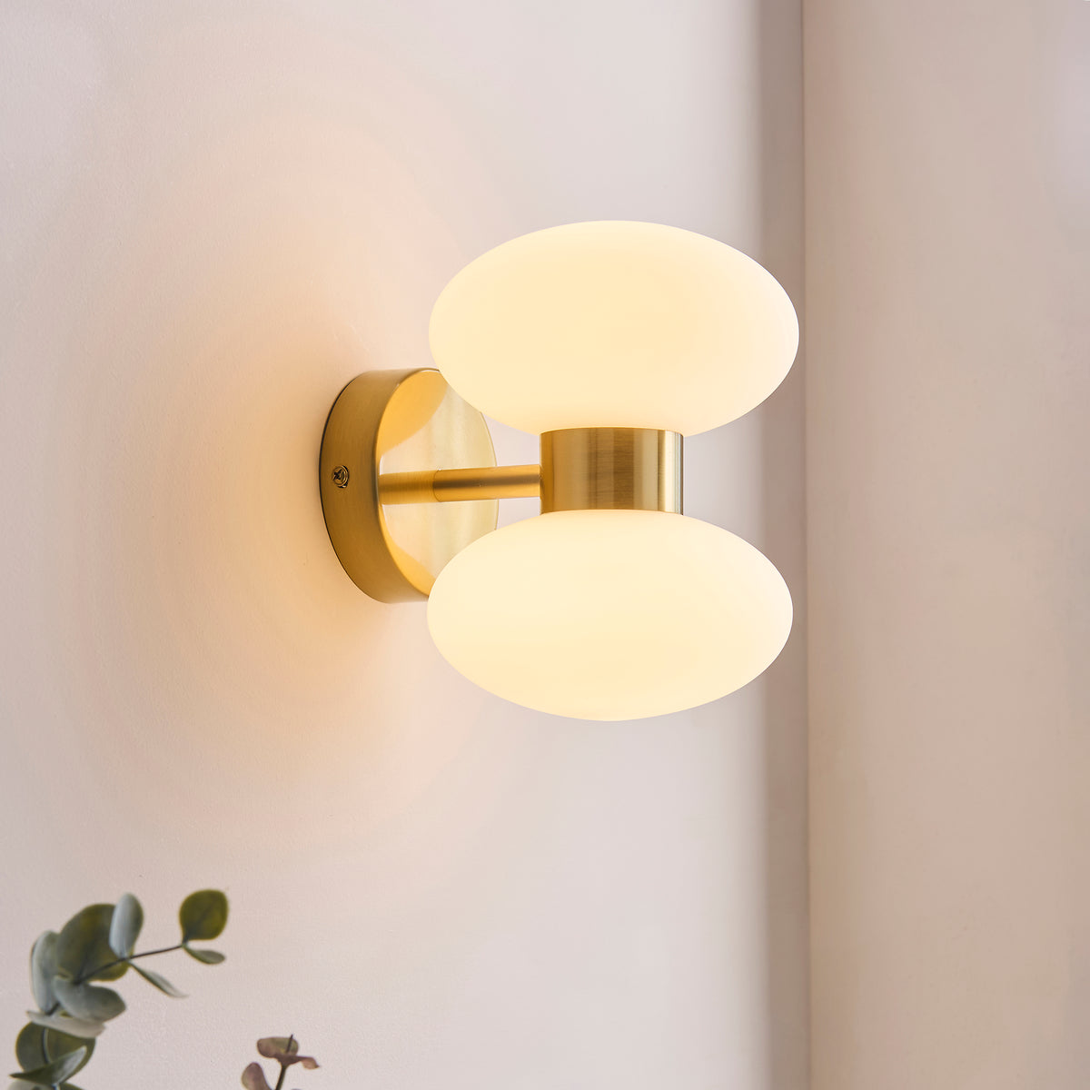 Lit lifestyle shot of the Endon Rossano 2 light wall light in warm brass with opal glass shades photographed straight on beside a potted plant