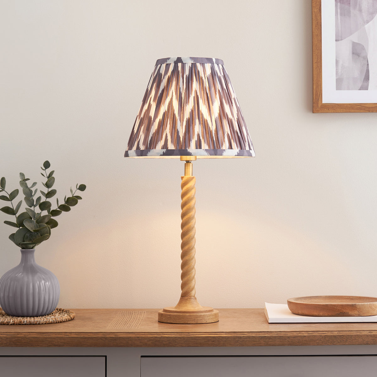 Endon Twist oak effect table lamp with grey zigzag shade lit on an oak sideboard beside a grey vase, eucalyptus and a framed print