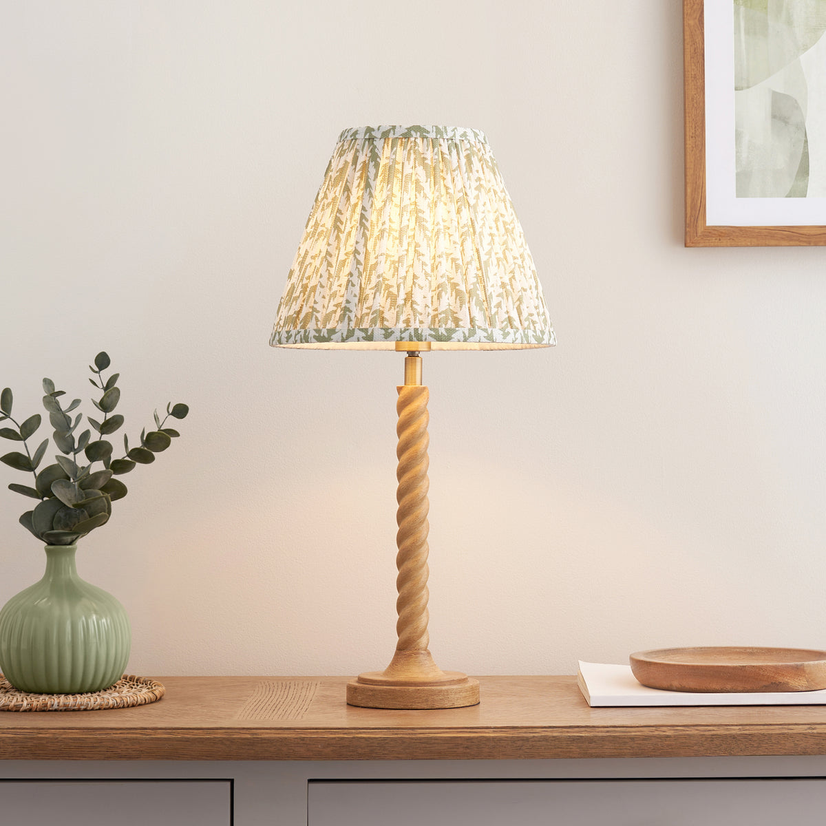 Endon Twist table lamp lit on an oak sideboard, oak effect twisted base with herb garden green leaf print shade, green vase and framed print alongside
