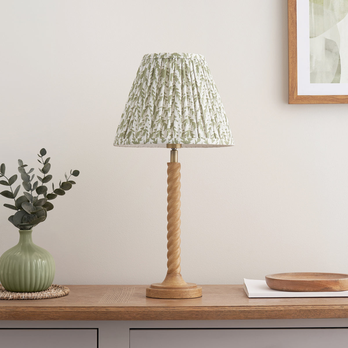 Endon Twist table lamp switched off on an oak sideboard, oak effect twisted stem and green leaf print pleated shade beside a green vase with eucalyptus