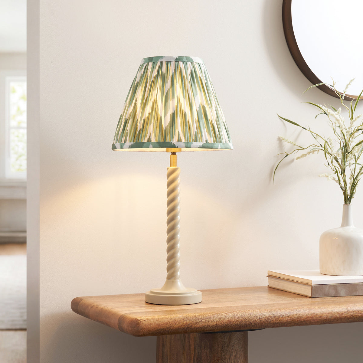 Endon Twist table lamp lit on a wooden console with a round mirror, white vase of dried grasses and books, almond white stem and green zigzag shade