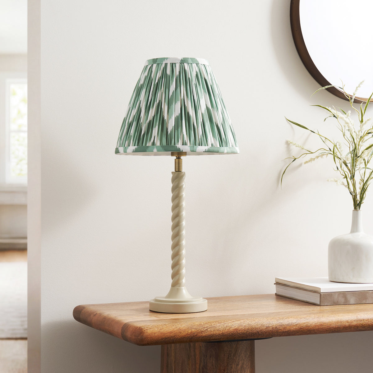 Endon Twist table lamp unlit on a wooden console beside a round mirror and white vase, almond white twisted stem, Cotswold green zigzag shade