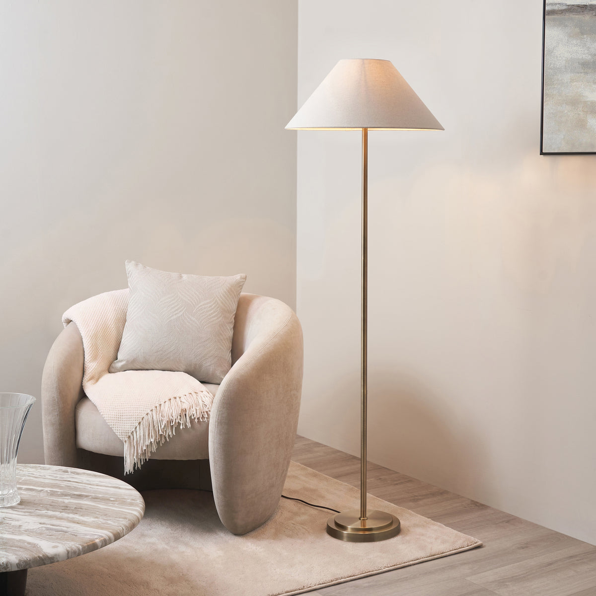Endon Arlo antique brass floor lamp with natural linen shade lit beside a cream armchair, marble side table and rug in a living room