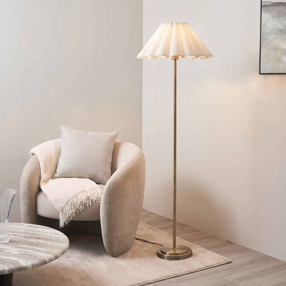 Endon Arlo antique brass floor lamp with vintage white scalloped shade, lit, standing beside a beige armchair, rug and marble side table