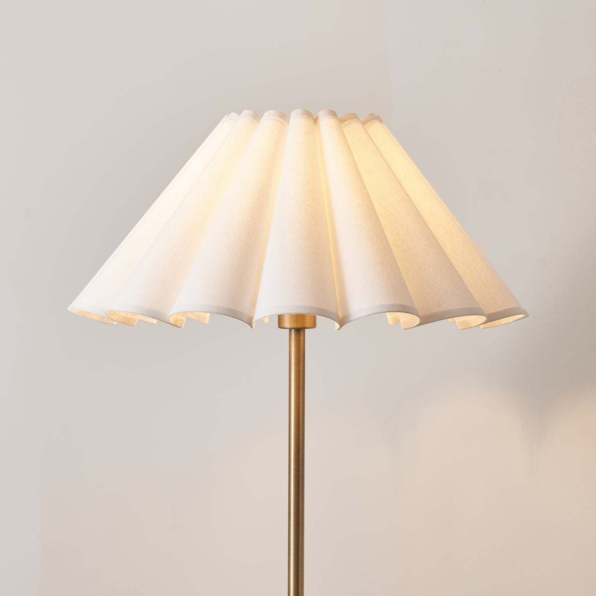 Close up of the lit Ayla 53cm scalloped vintage white cotton shade on the Endon Arlo antique brass floor lamp stem