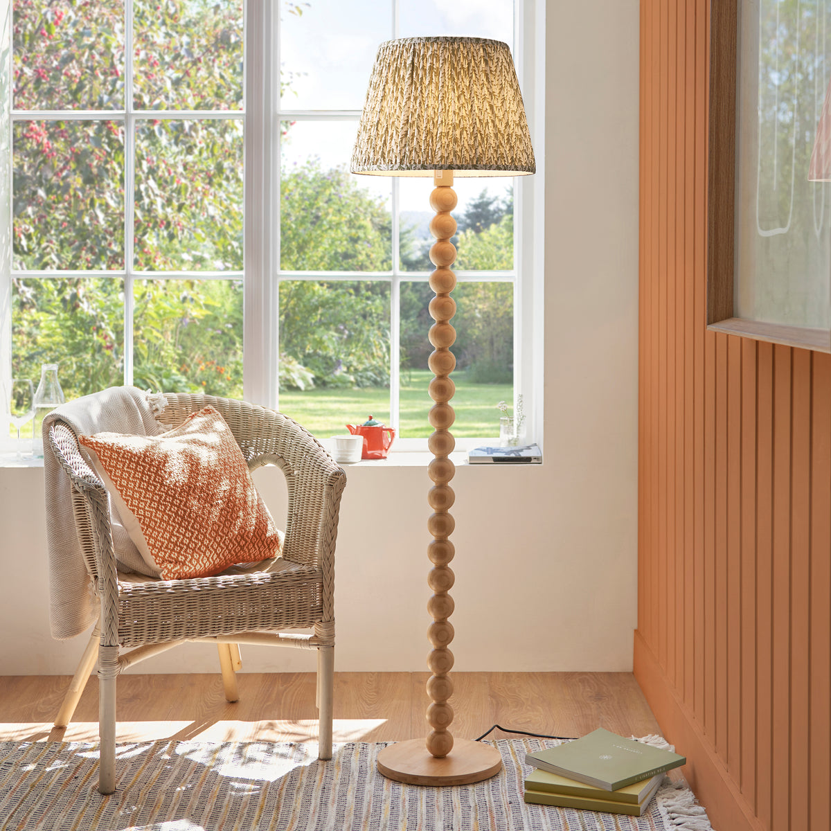 Endon Cane bamboo floor lamp lit in a sunny living room beside a wicker armchair with an orange cushion, green leaf shade glowing by a window