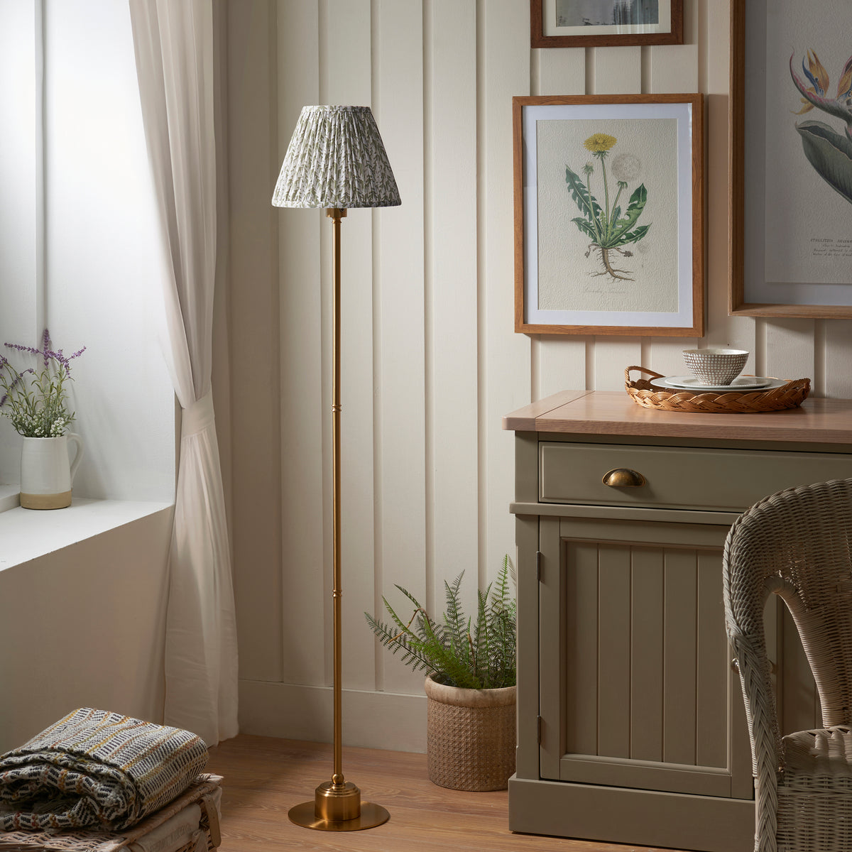 Endon Burley brass floor lamp unlit with green leaf shade, standing beside a painted sideboard and a window with lavender in a cottage room