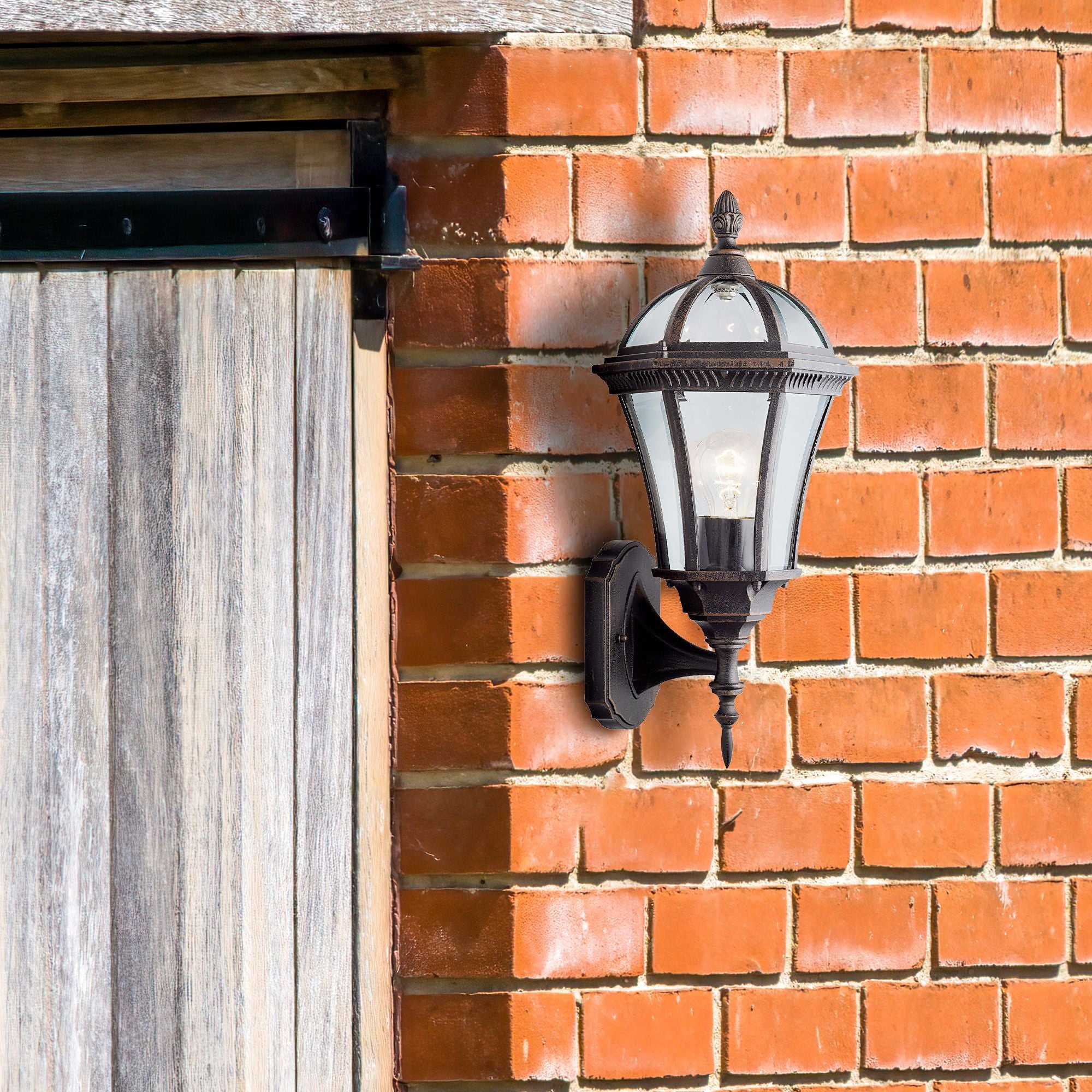 Capri Outdoor Uplight mounted on a red brick wall beside a wooden barn door, lit with a warm glow