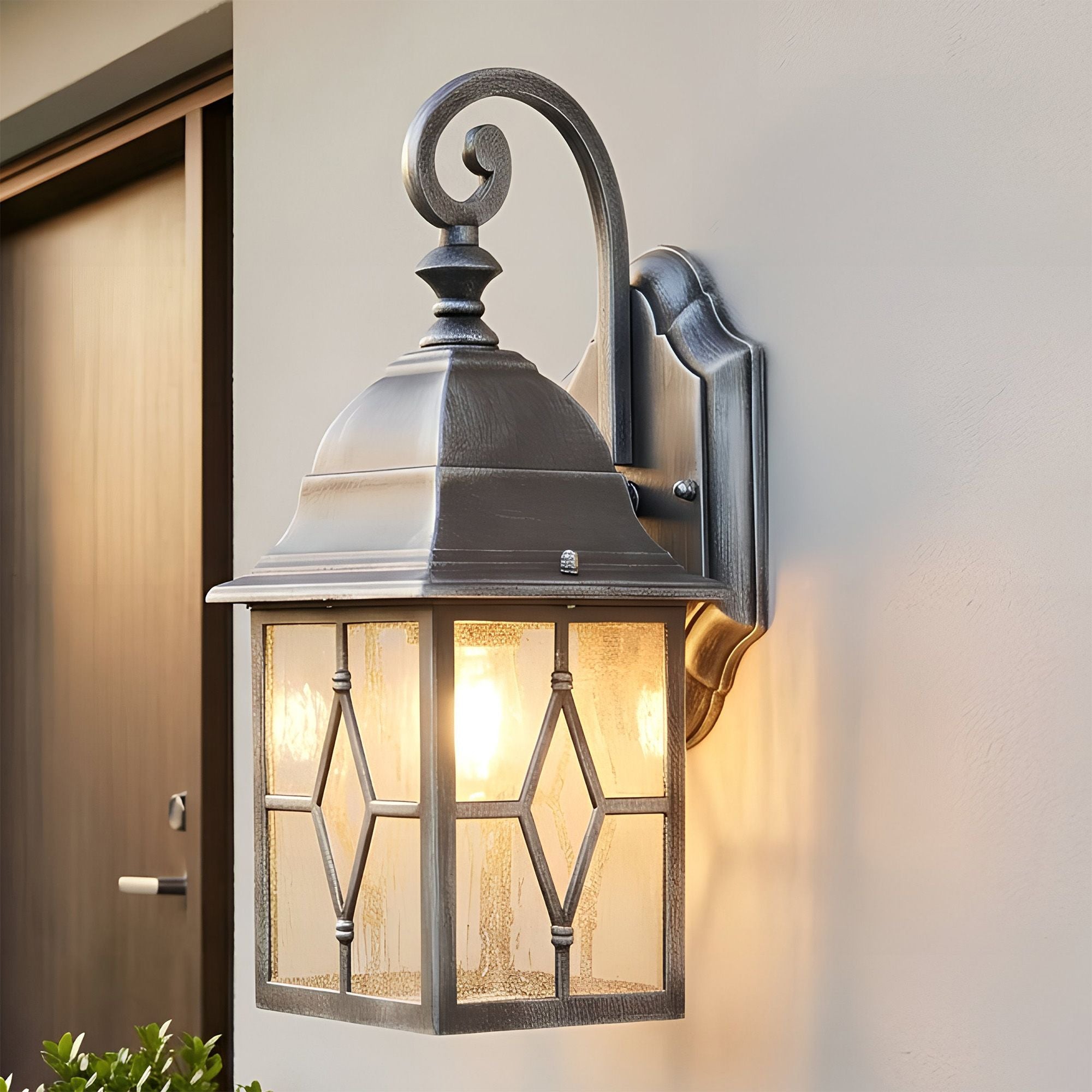 Genoa outdoor wall light mounted beside a front door, lit warmly at dusk with greenery in the foreground