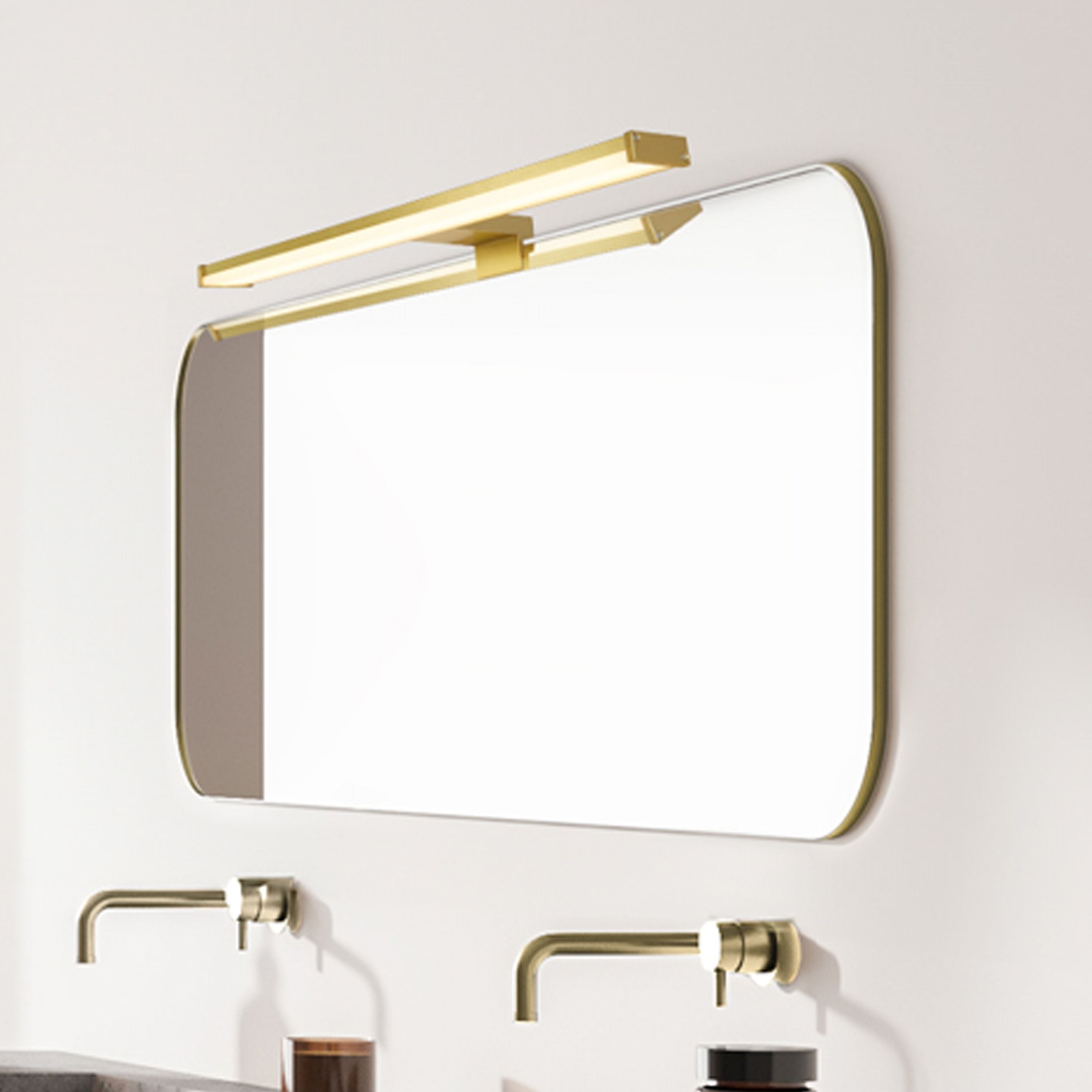 Searchlight Kos LED bathroom mirror light in gold mounted above a rounded rectangular mirror in a bathroom, with brass tap fixtures below