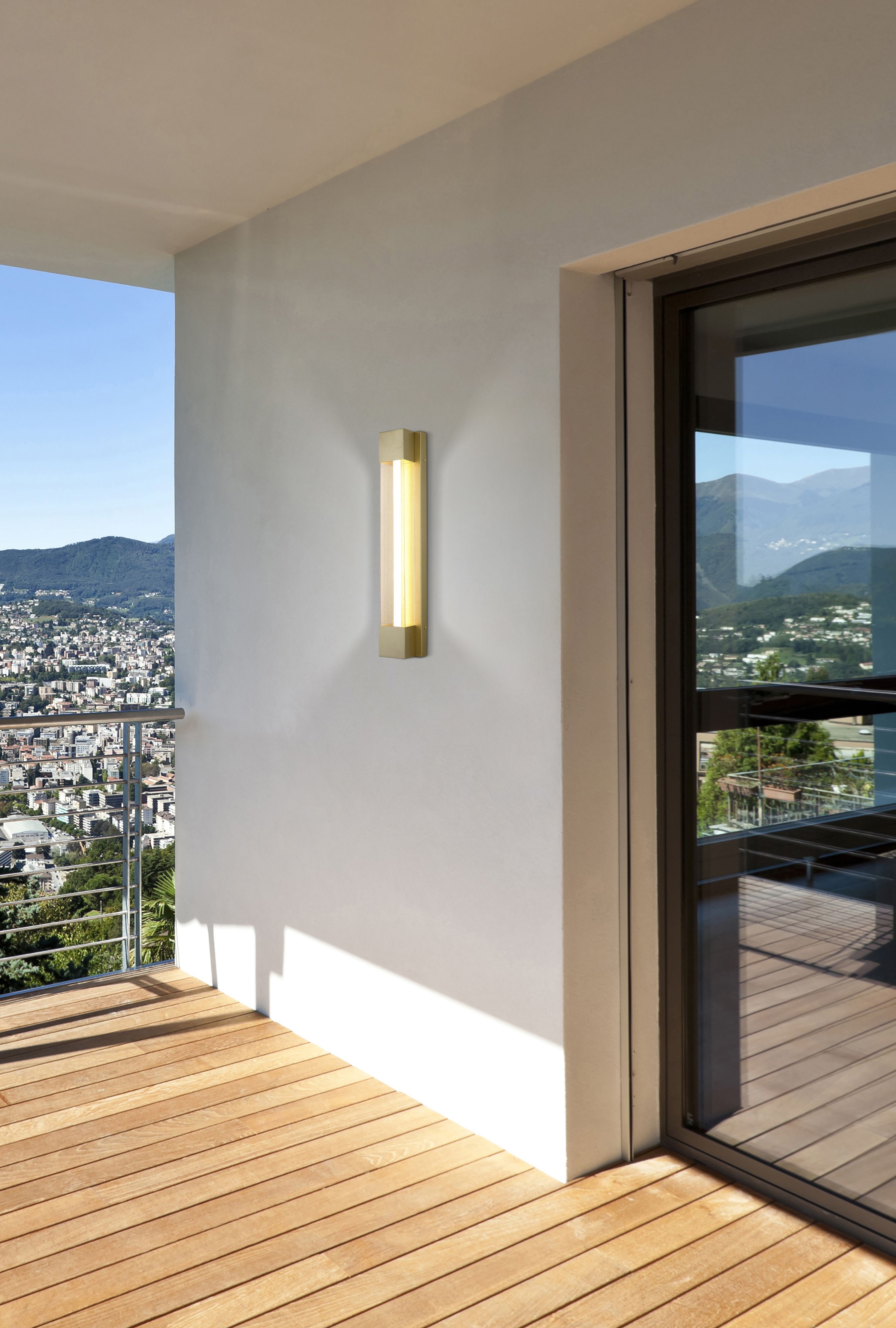 Sacramento 60cm outdoor LED wall light in painted gold installed on an exterior wall beside a glazed door, lit at dusk with a city view in the background
