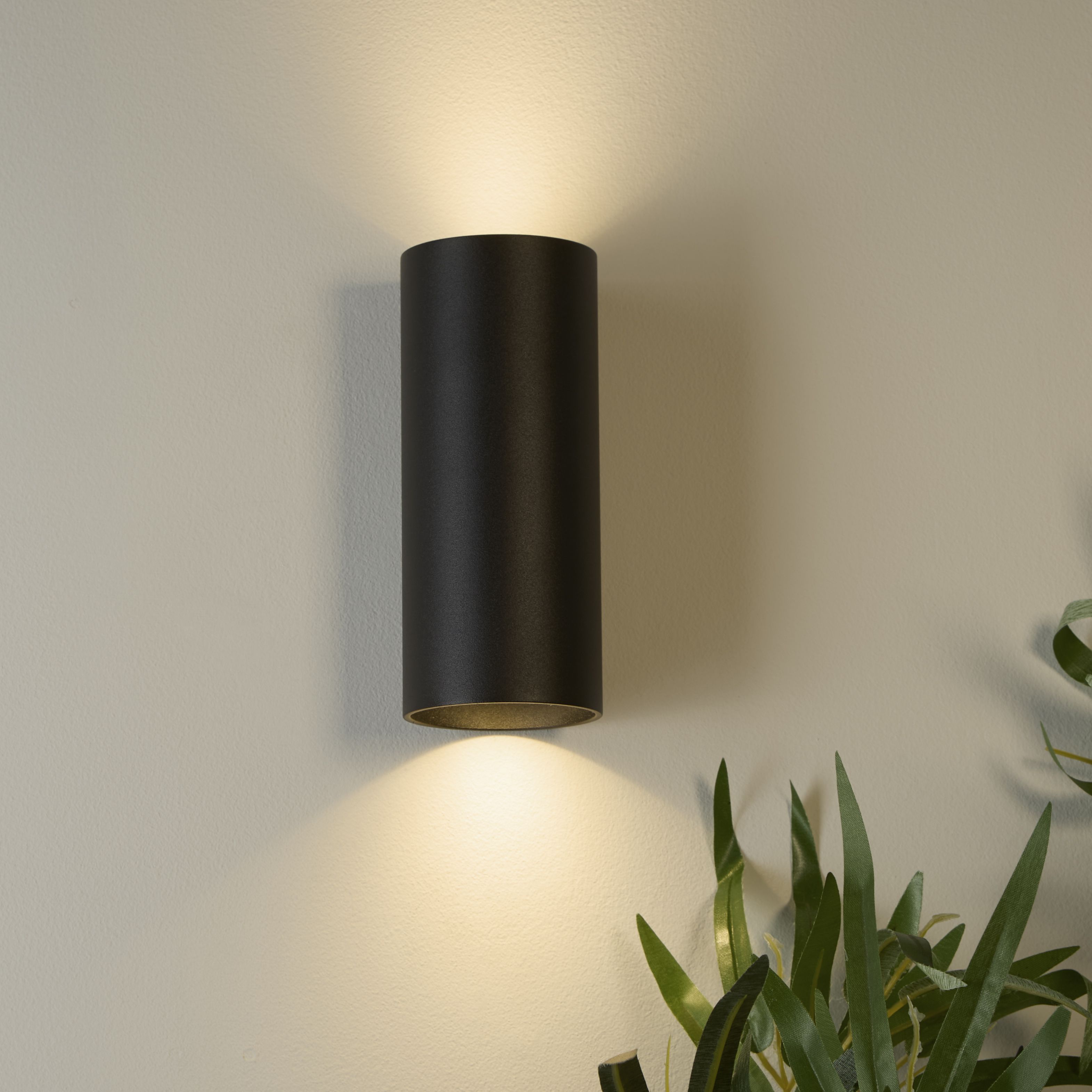 Searchlight Cyls wall light in matte black mounted on a warm toned wall beside a leafy plant, lit both upward and downward