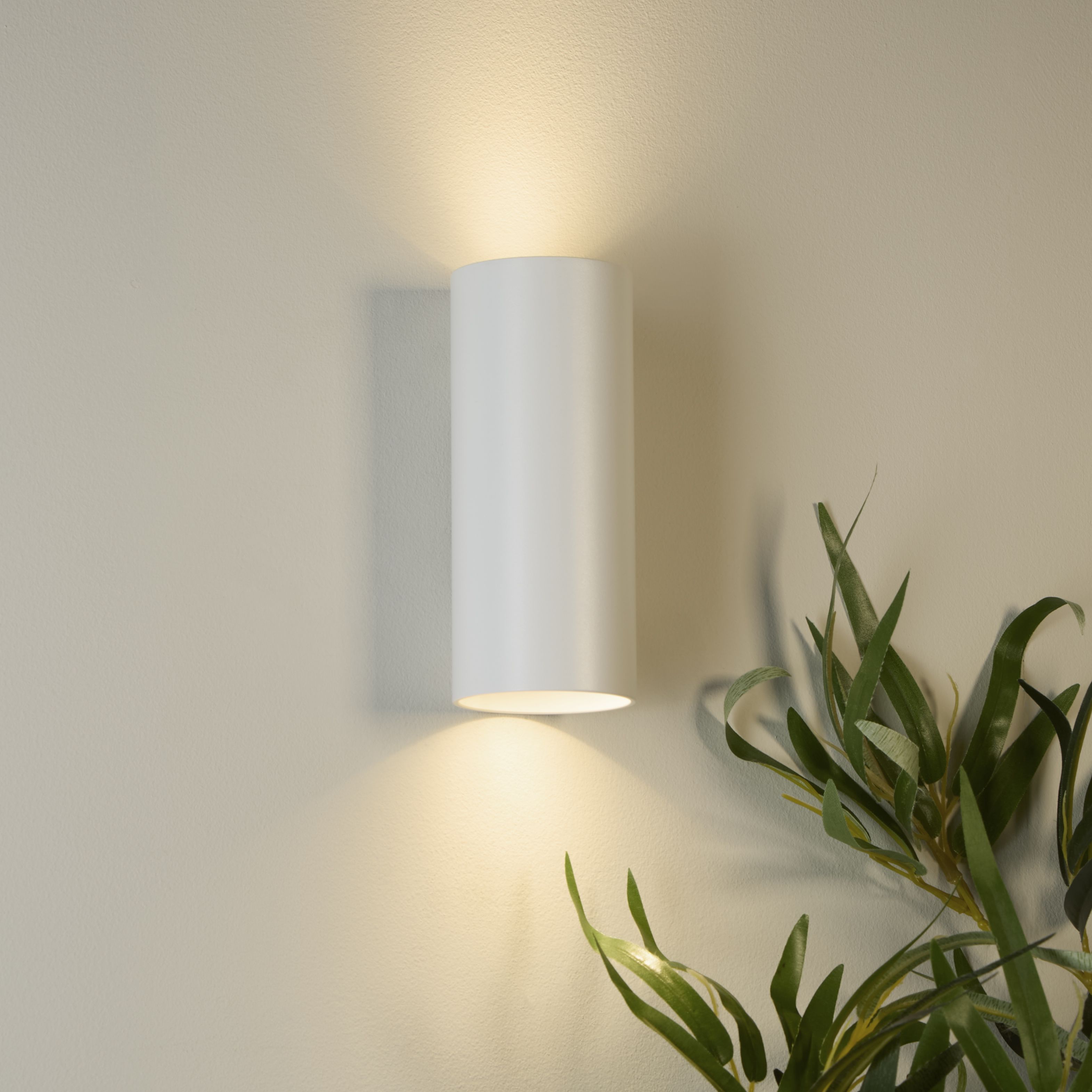 Searchlight Cyls wall light in white mounted on a warm toned wall beside a leafy plant, lit both upward and downward