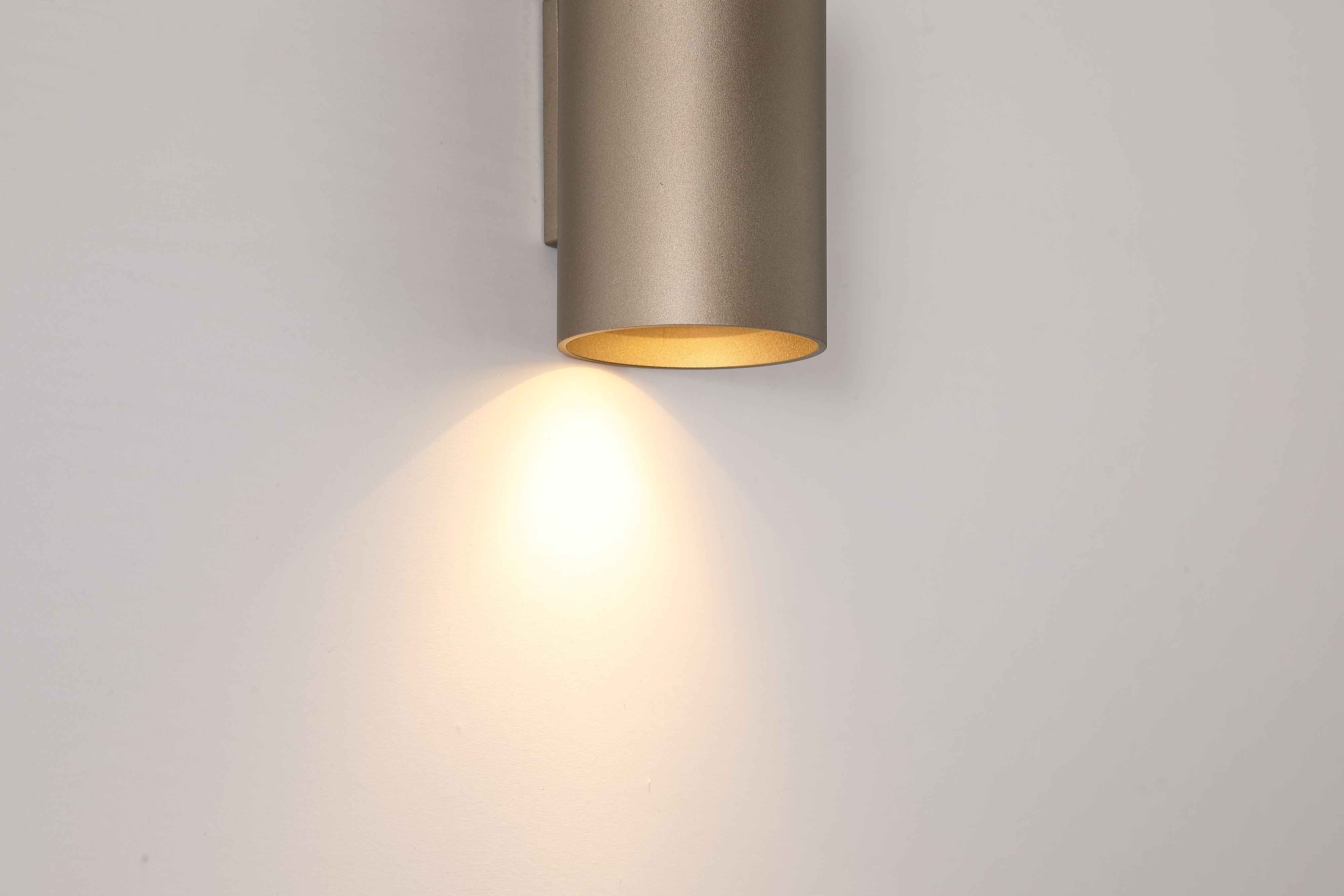 Close up of the Searchlight Cyls Cylinder Down Wall Light in latte, lit, showing the warm gold interior of the shade and the light beam falling on the wall