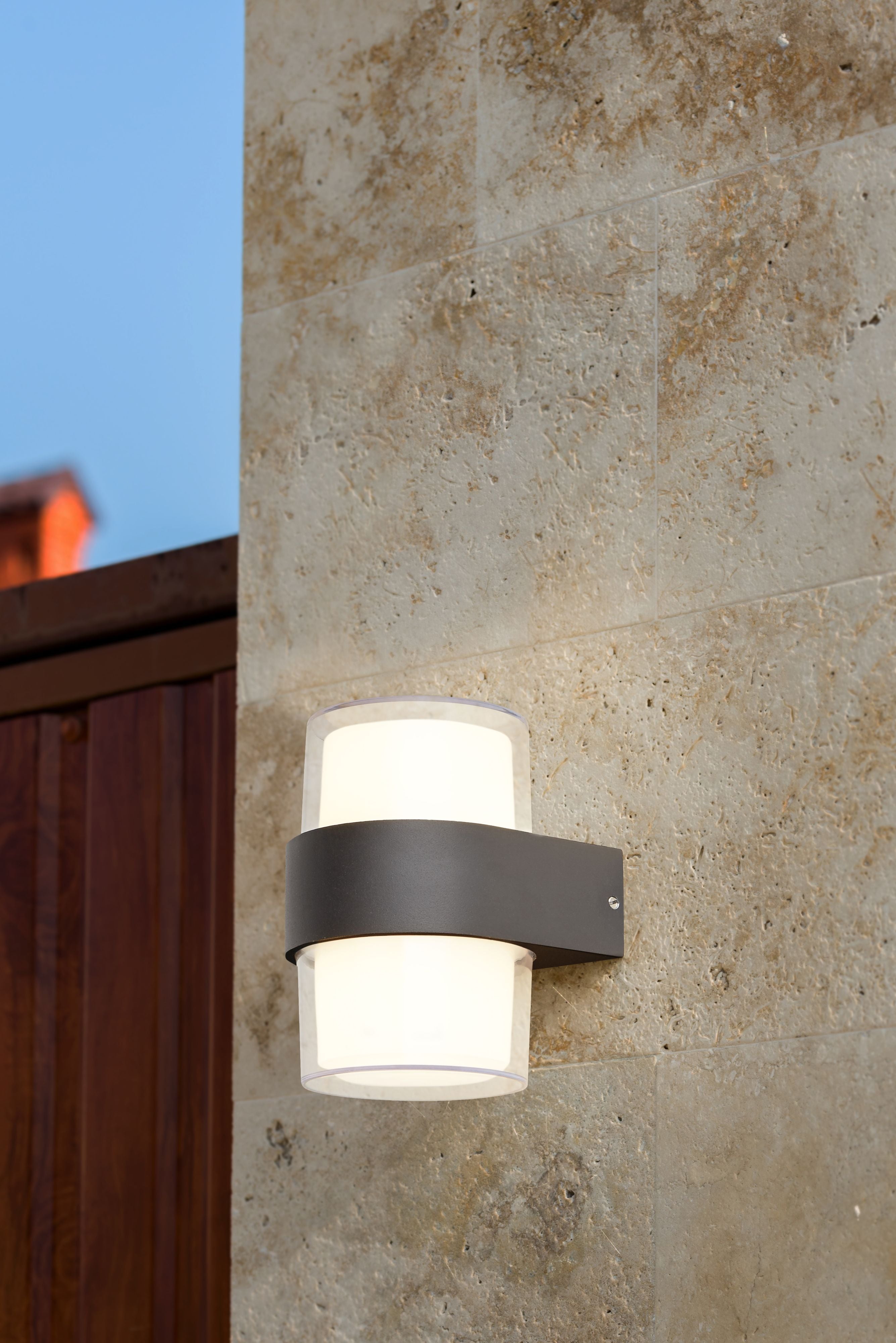 Searchlight Pittsburgh round outdoor wall light installed on a stone wall beside a wooden gate, lit warm white above and below the black band at dusk