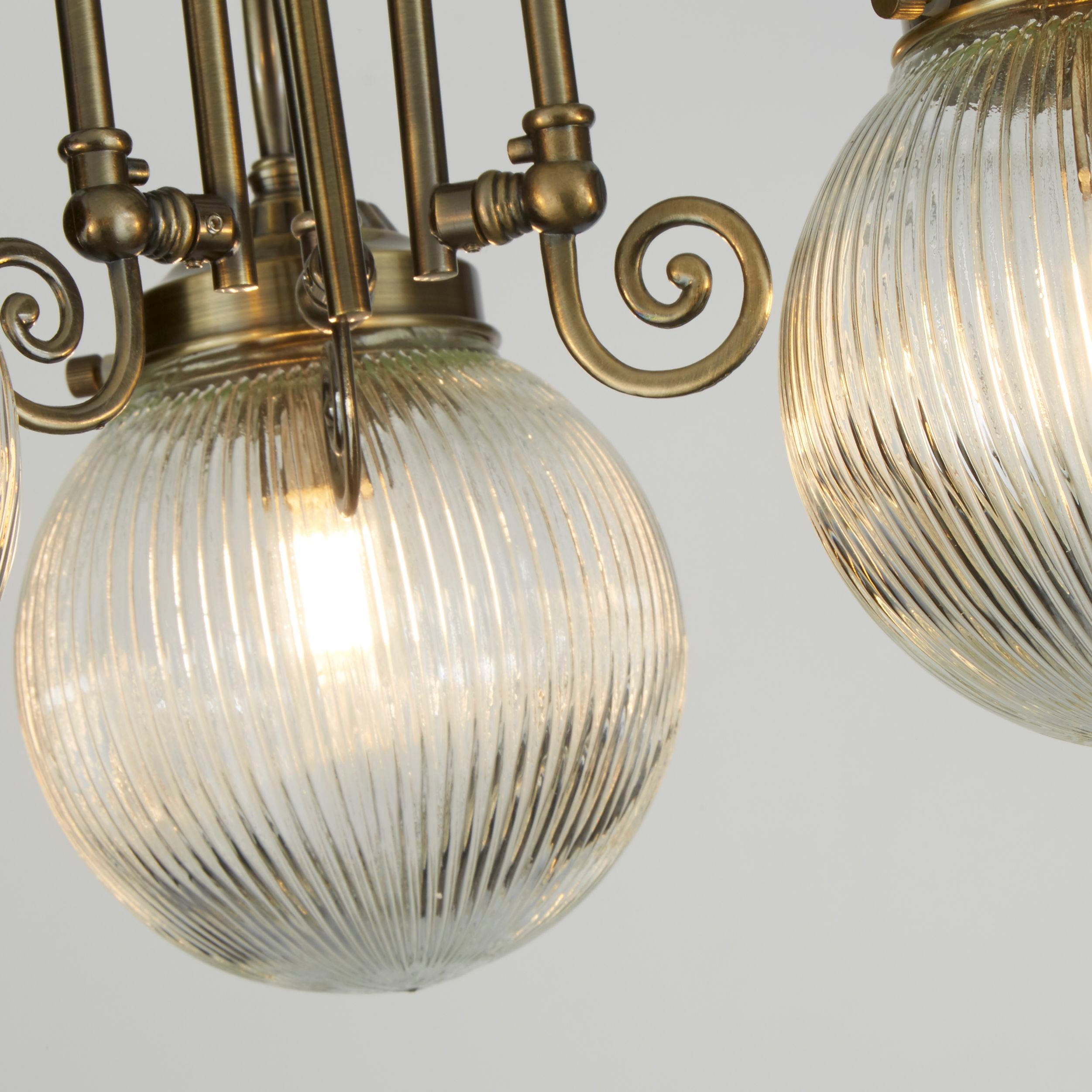 Close up of one ribbed glass globe on the Belvue flush light, lit warm white, showing the antique brass gallery and scrollwork