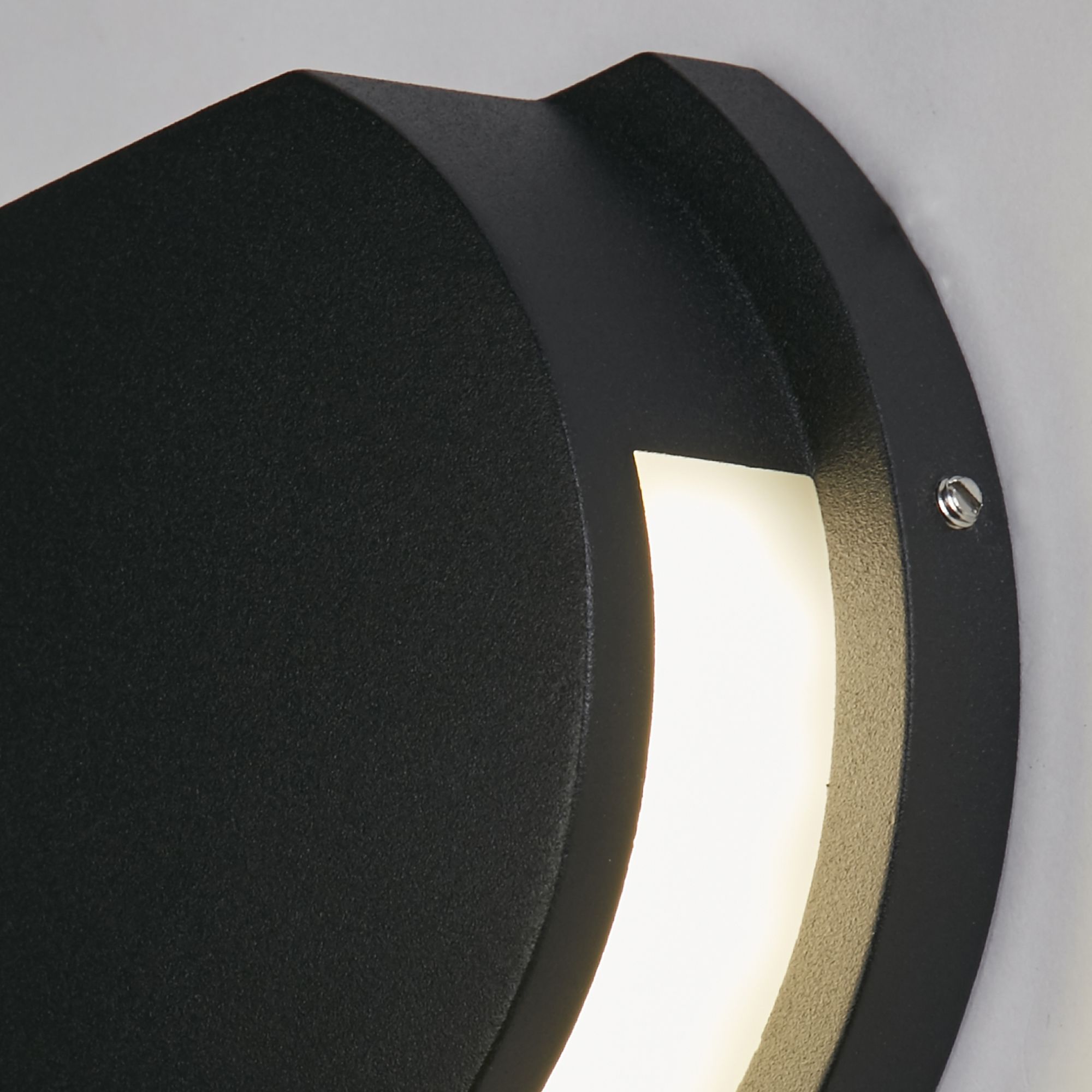 Close up of the Searchlight Chiswick LED outdoor wall light showing the matte black finish and the lit crescent shaped acrylic diffuser with a visible fixing screw