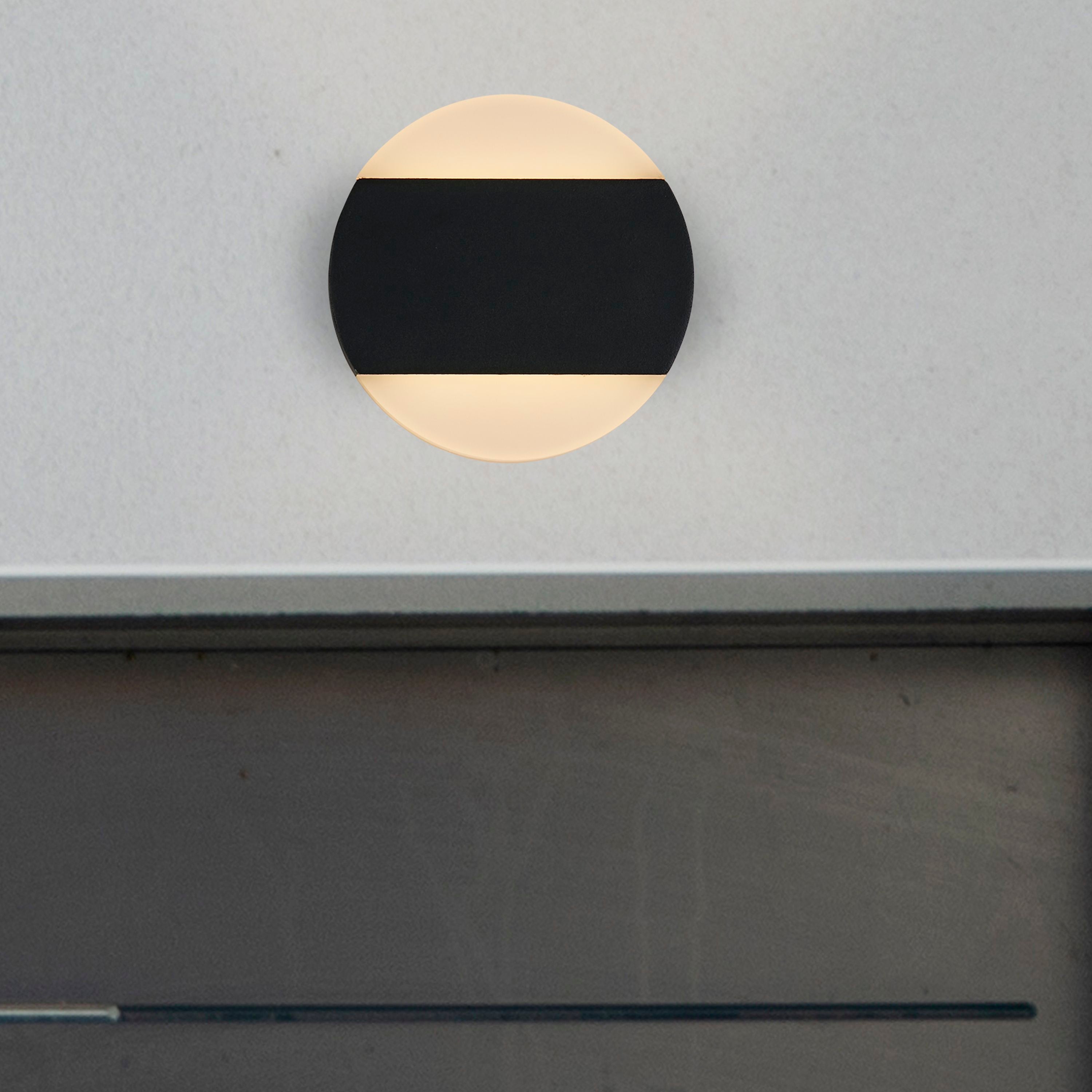 Searchlight Cavern LED outdoor wall light installed on a grey rendered exterior wall, lit warm white above and below the black band