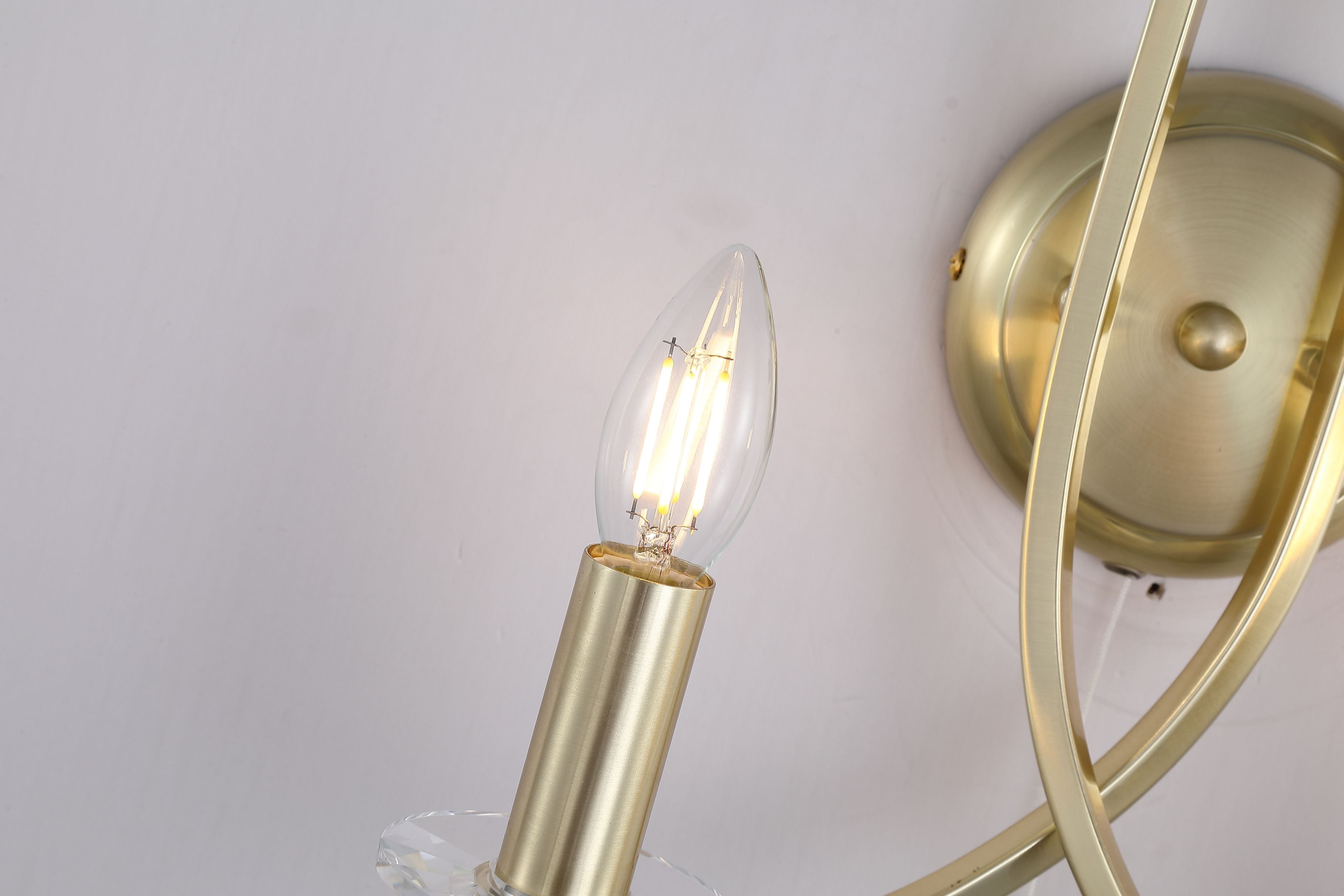 Close up of the Ascona 2 Light Wall Light showing a lit candle bulb, crystal style sconce and satin brass wall plate with crossed arms
