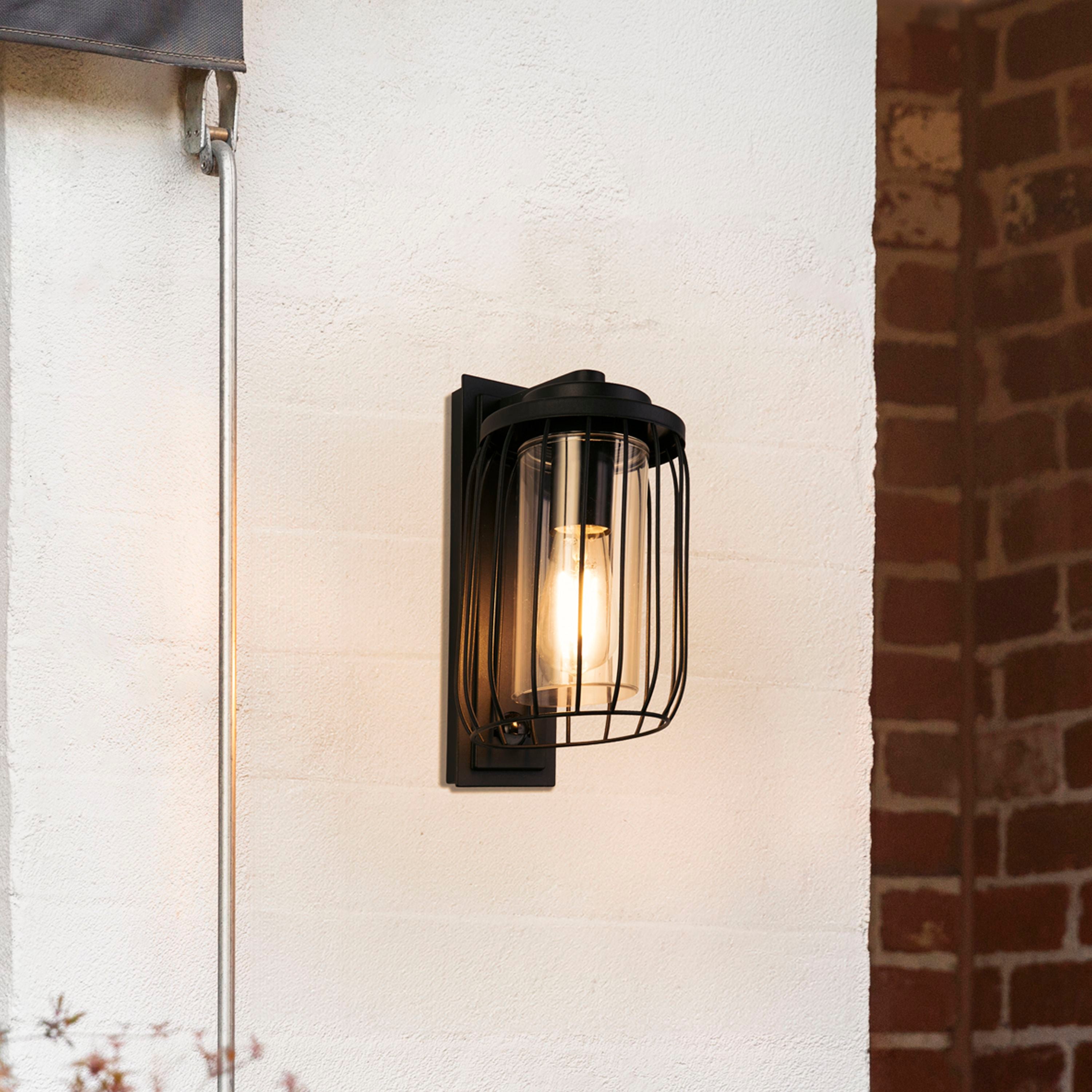 Reed 1 Light Wall Light with PIR mounted on a rendered exterior wall beside a brick pillar, lit at dusk next to a rolled up awning.