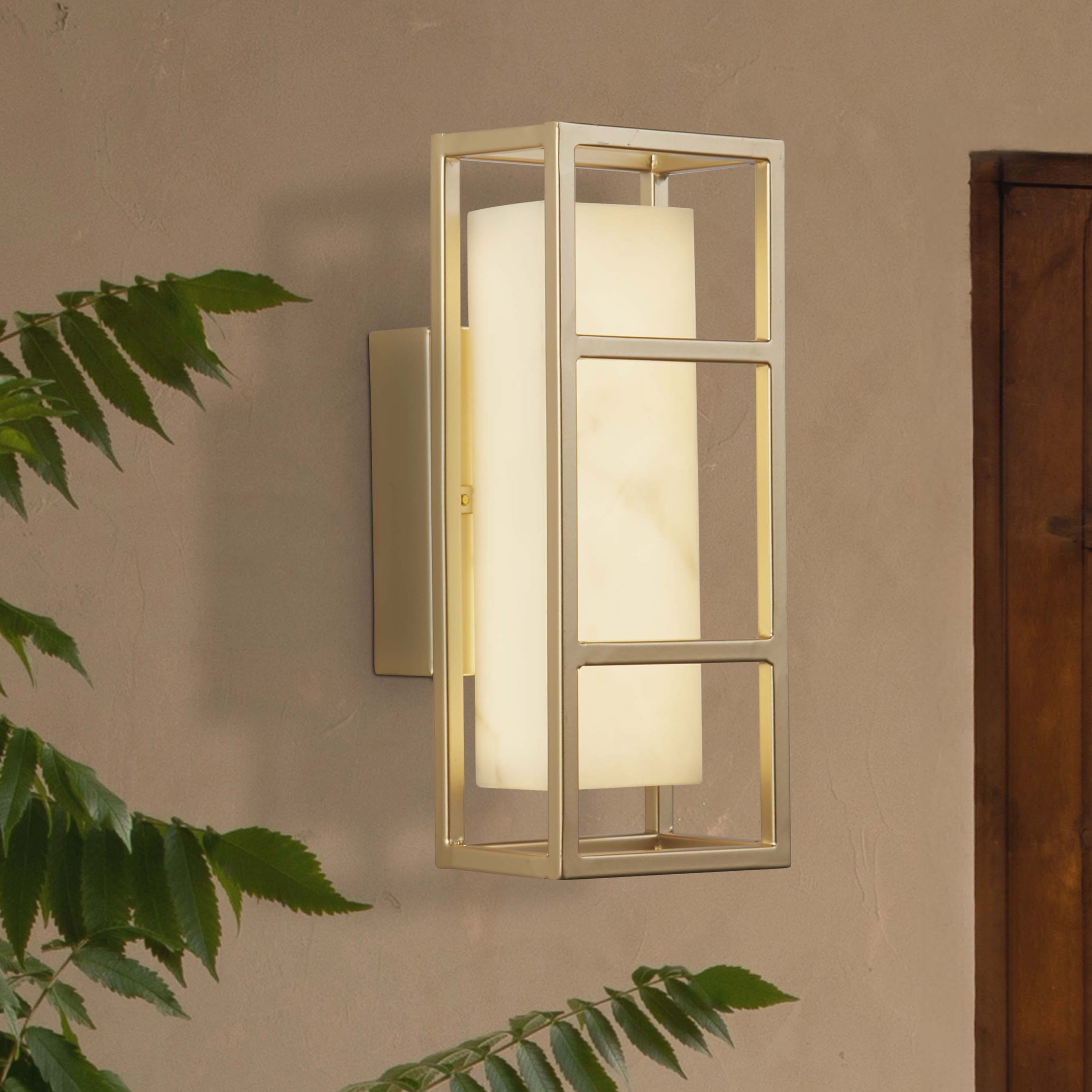 Searchlight Mirage LED wall light in matt gold, styled lifestyle shot mounted on a textured wall beside a door, with trailing green foliage in frame