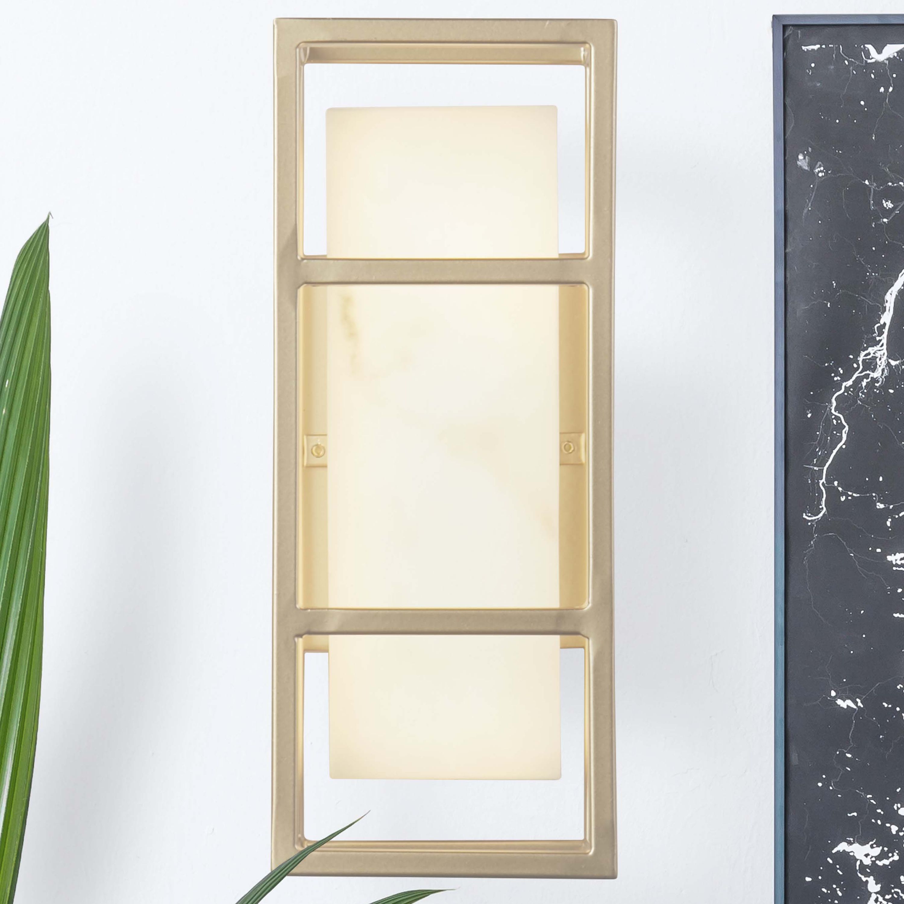 Searchlight Mirage LED wall light in matt gold mounted on a white indoor wall next to a framed black marble artwork, styled with a potted palm plant