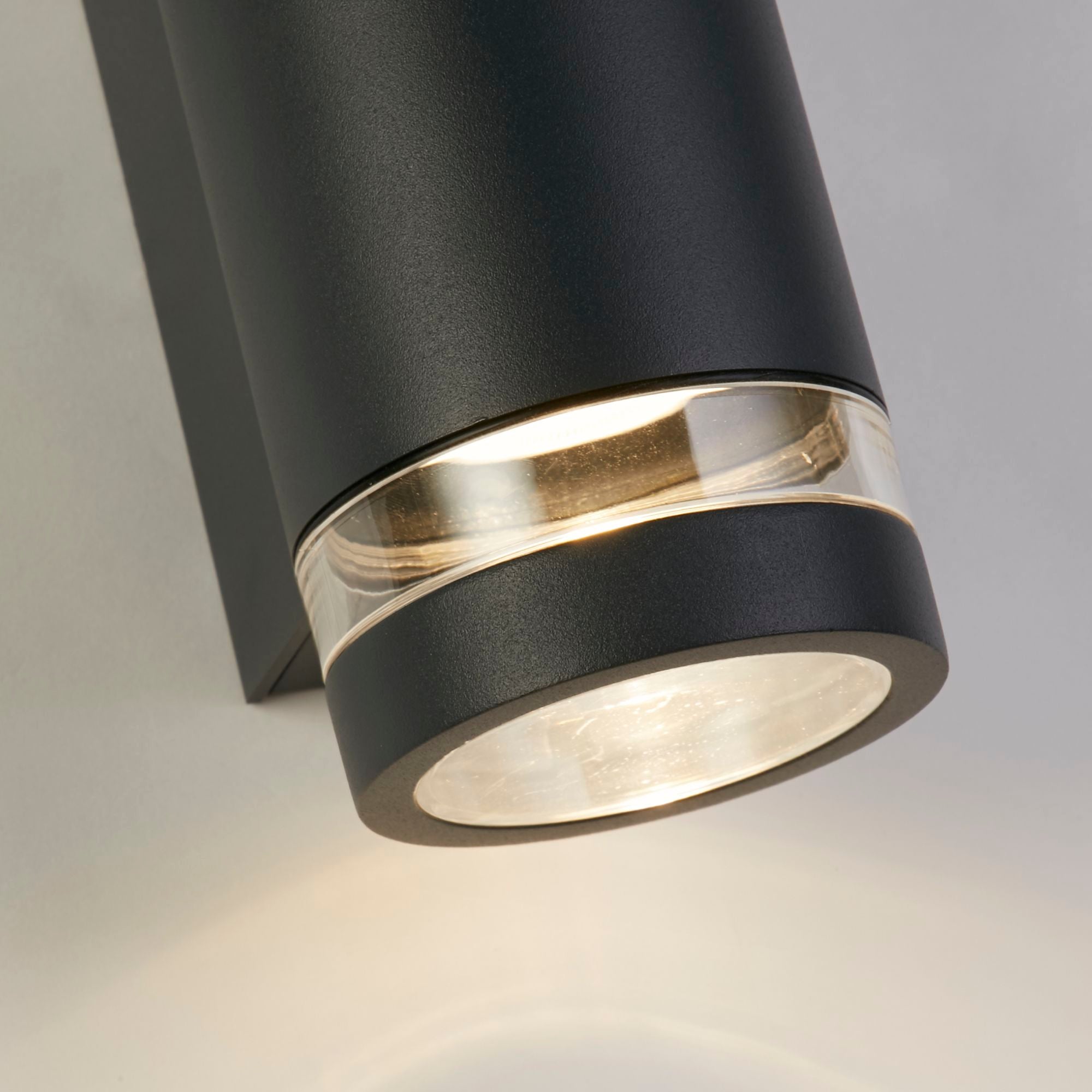 Close up angled view of the illuminated lower lens ring on the Austin grey wall light, mounted on a white wall