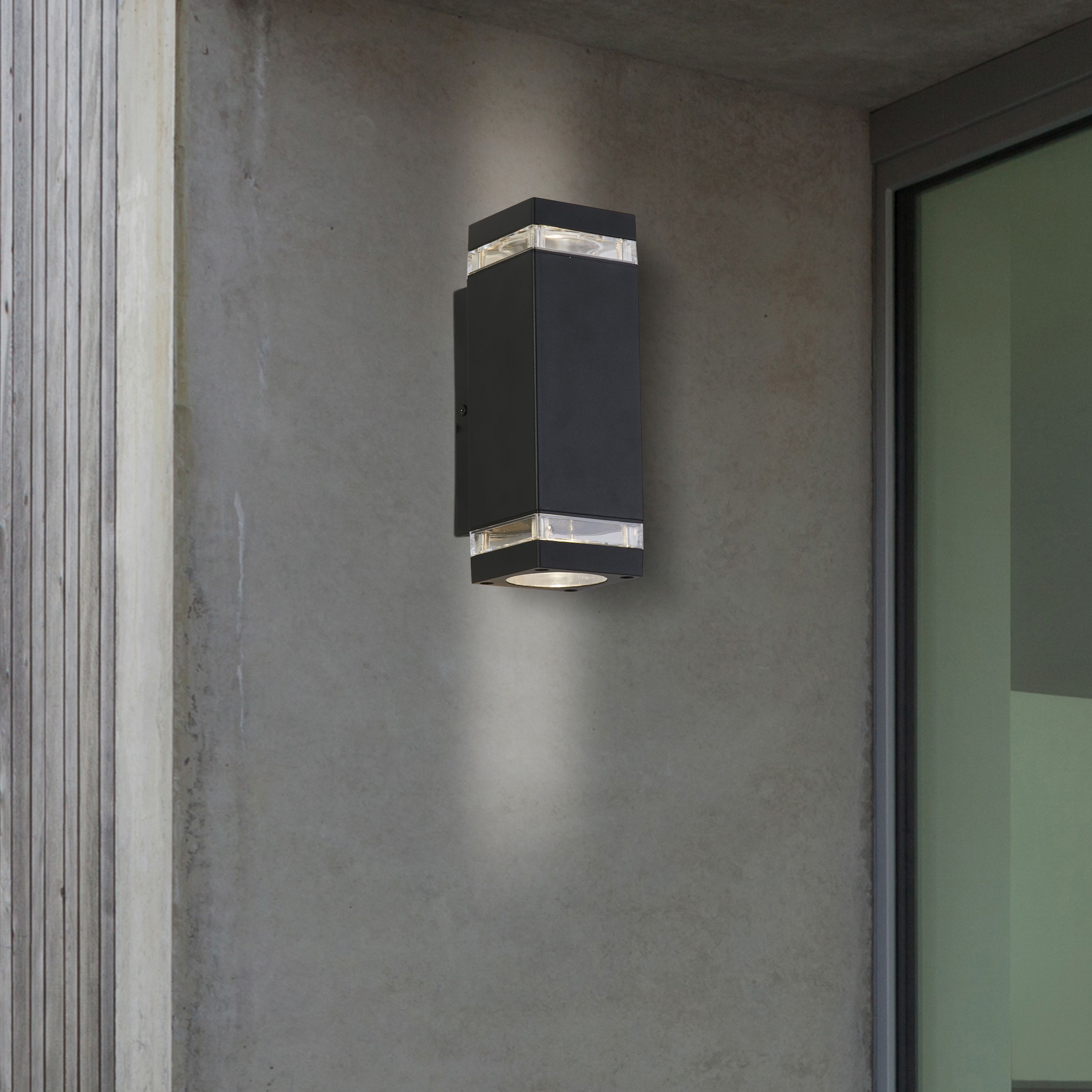 Searchlight Houston black outdoor wall light installed beside a front door on a textured concrete wall, lit at night with light spilling up and down