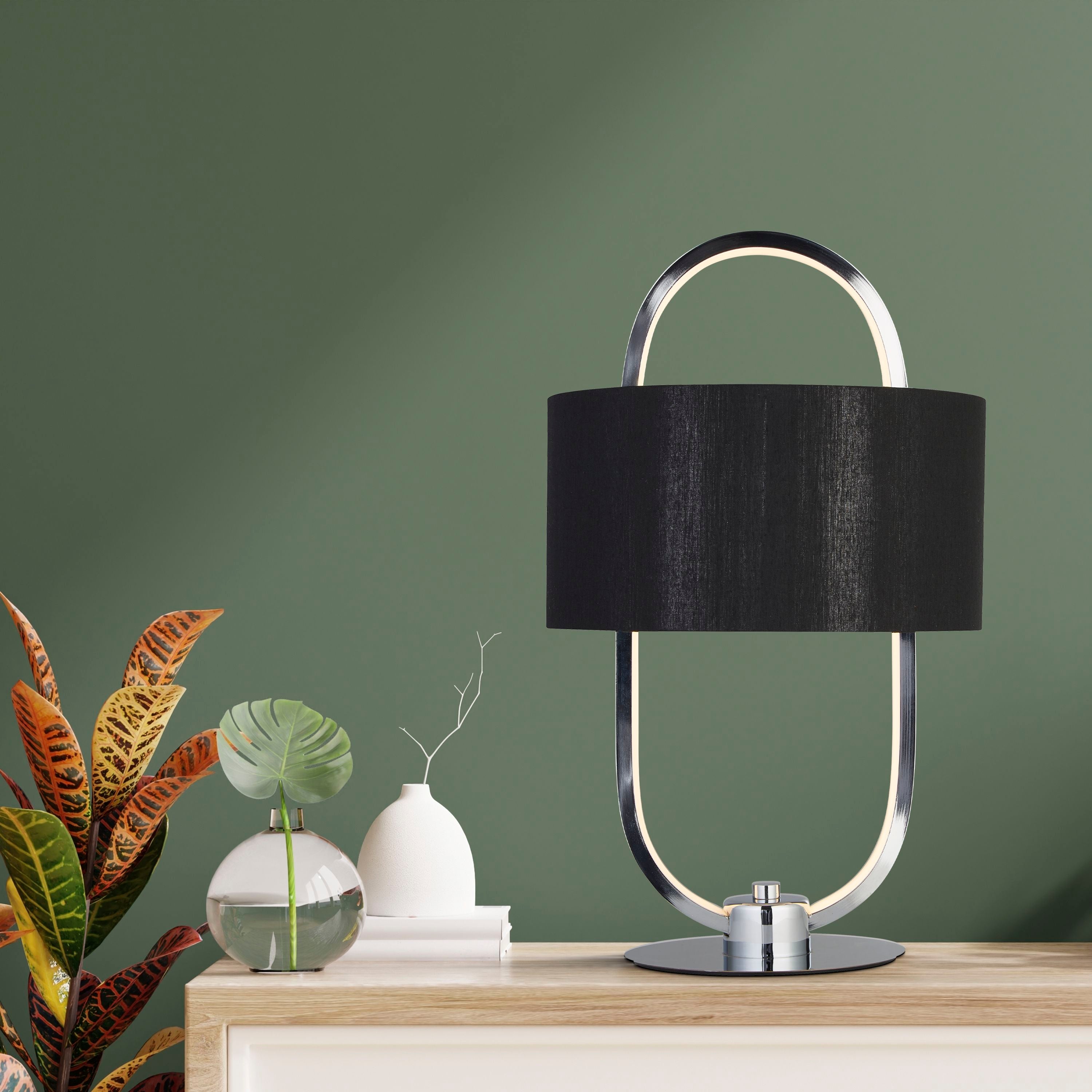 Madrid table lamp switched on and glowing on a light wood sideboard against a sage green wall with plants