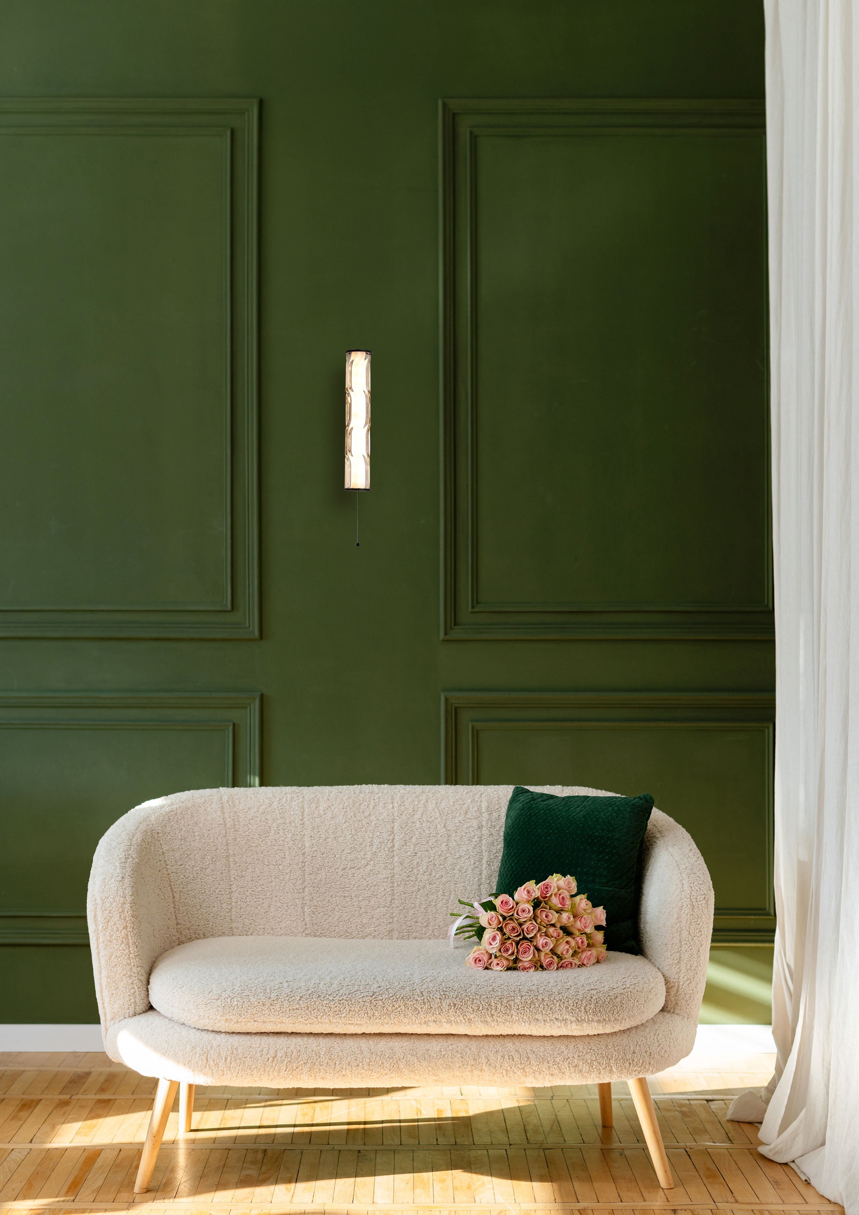 Matt black Vibrate wall light mounted on a dark green panelled wall beside a cream boucle sofa in a styled living room