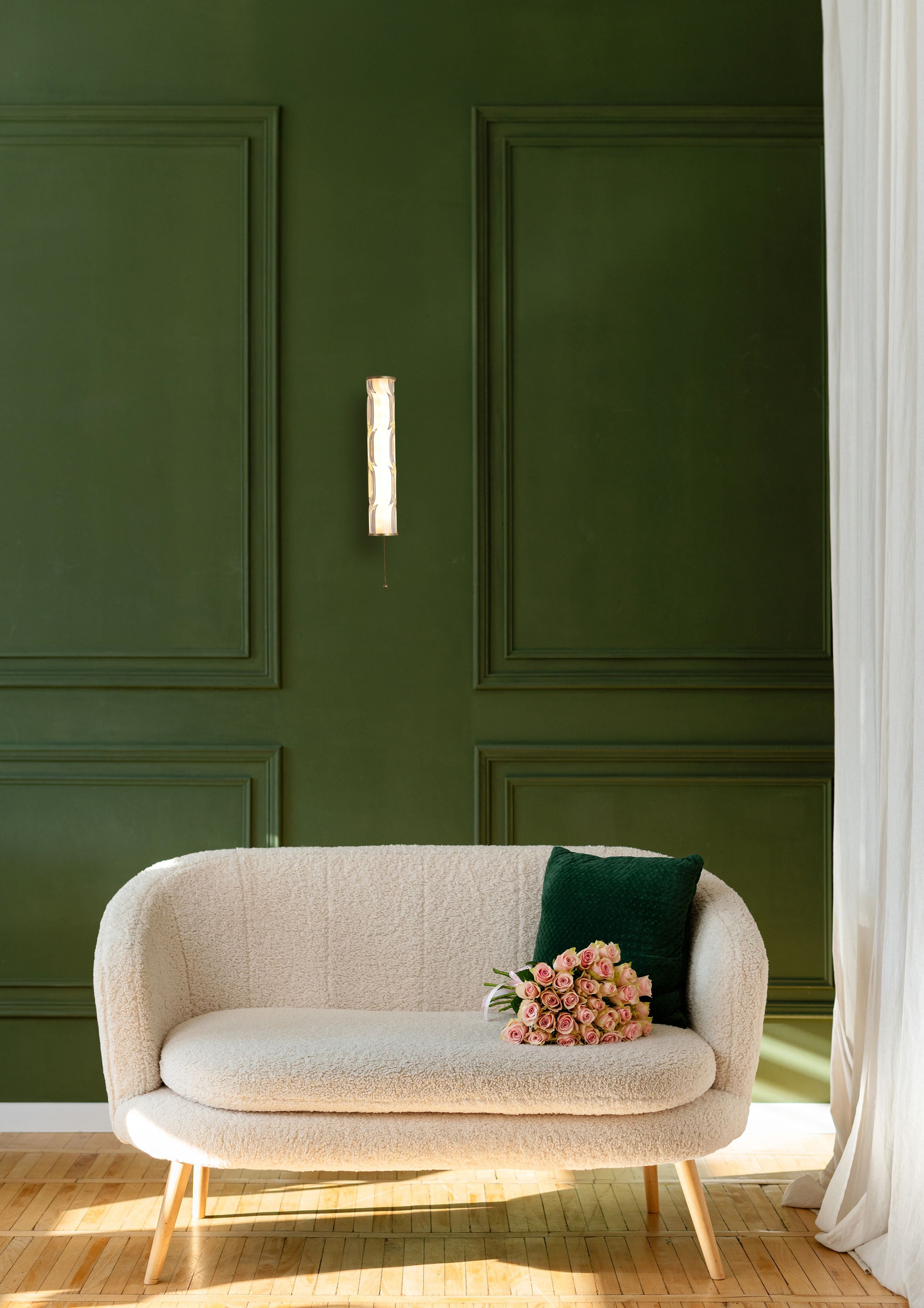Vibrate LED wall light in matt gold styled on a dark green panelled wall above a cream boucle sofa with a green cushion and pink roses