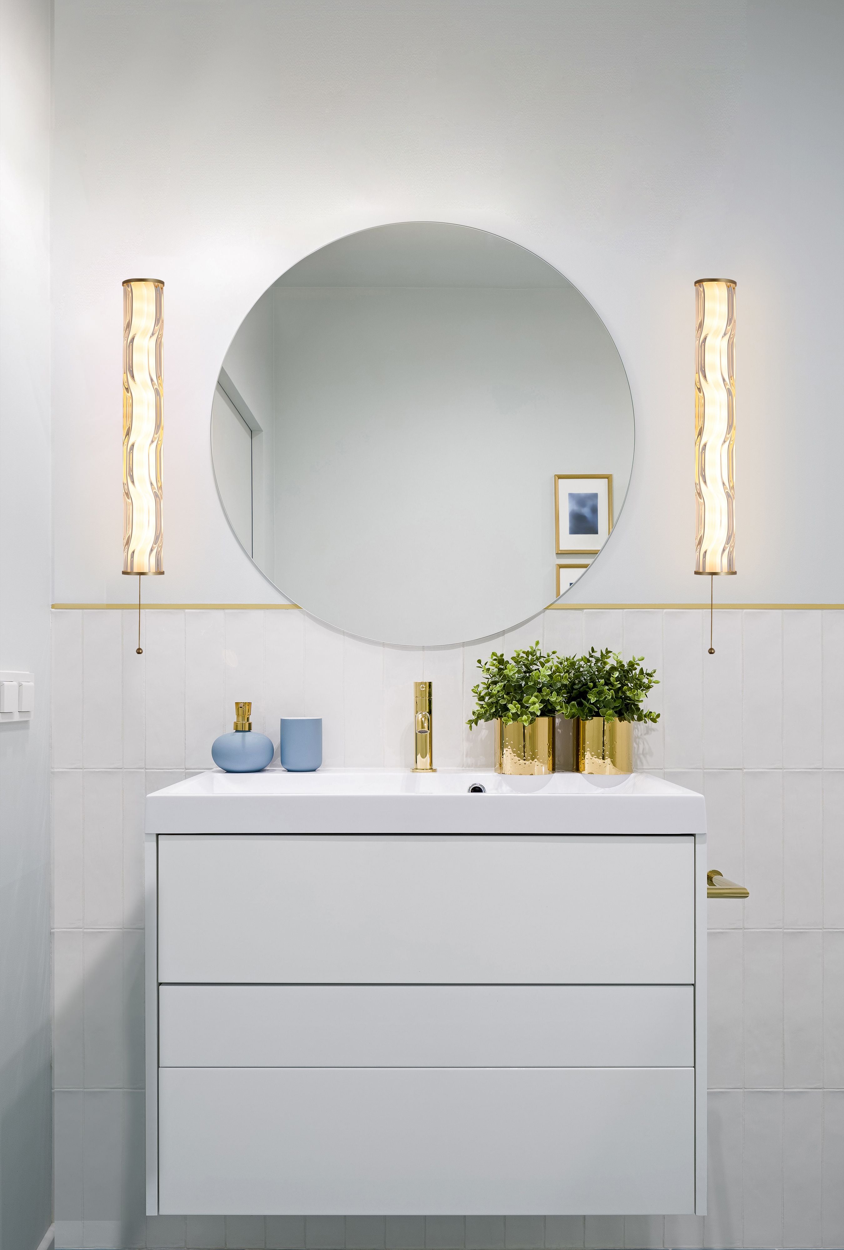 Pair of Vibrate LED wall lights in matt gold mounted either side of a round mirror above a white bathroom vanity with gold tapware