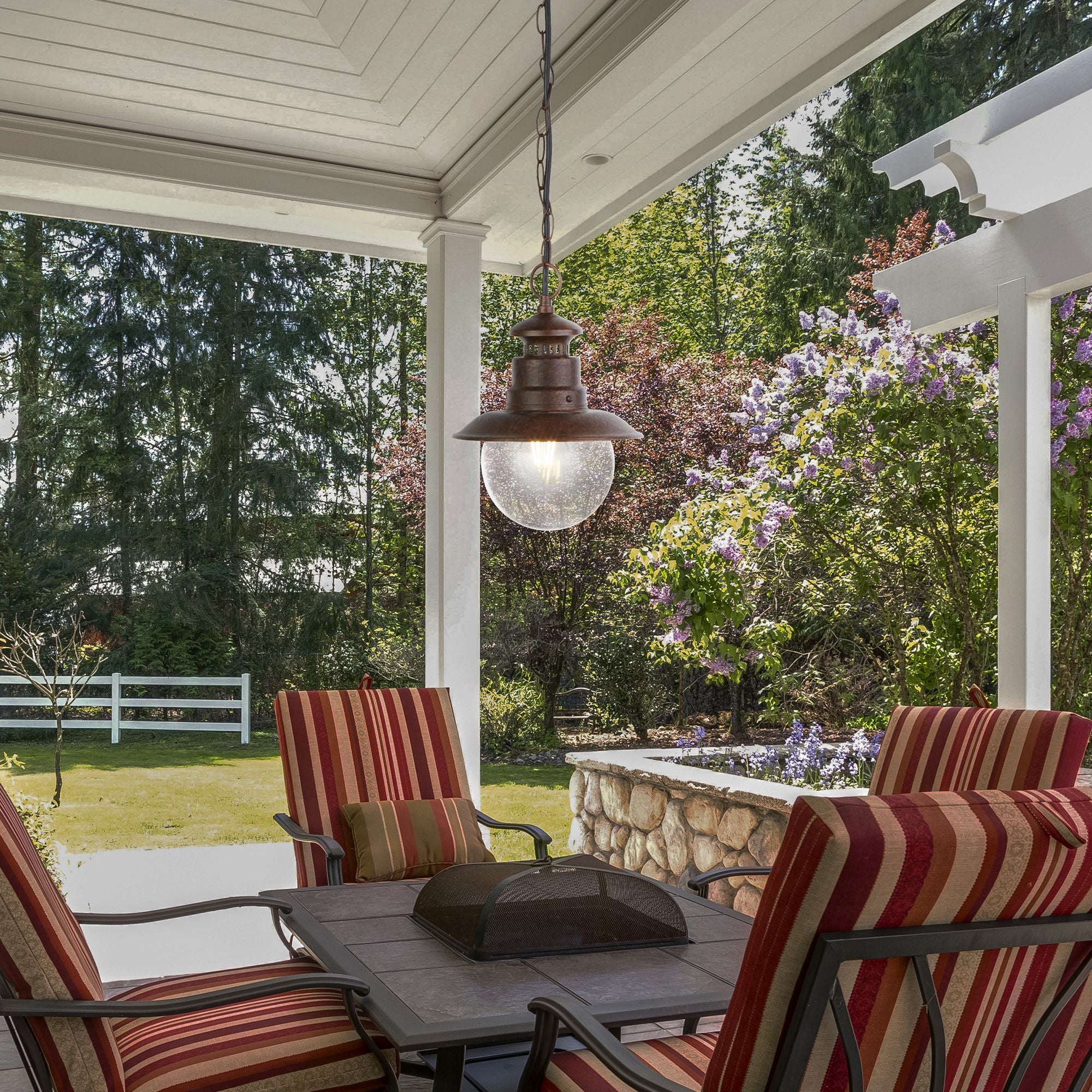 Station outdoor pendant hanging lit above a table on a covered porch furnished with striped cushioned chairs and a garden view behind