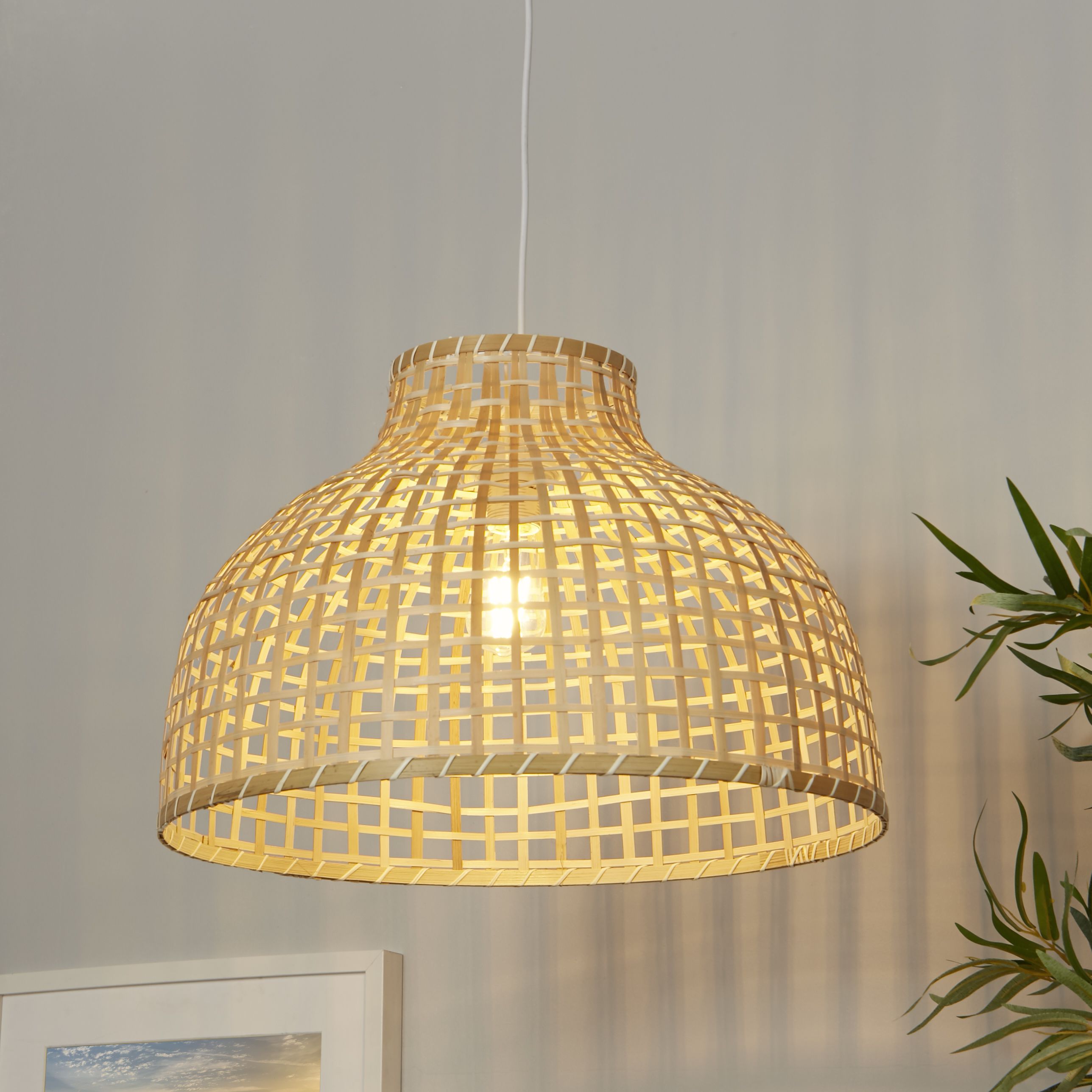 Bali Pendant in bamboo hanging in a styled room, illuminated against a grey wall and beside trailing green foliage