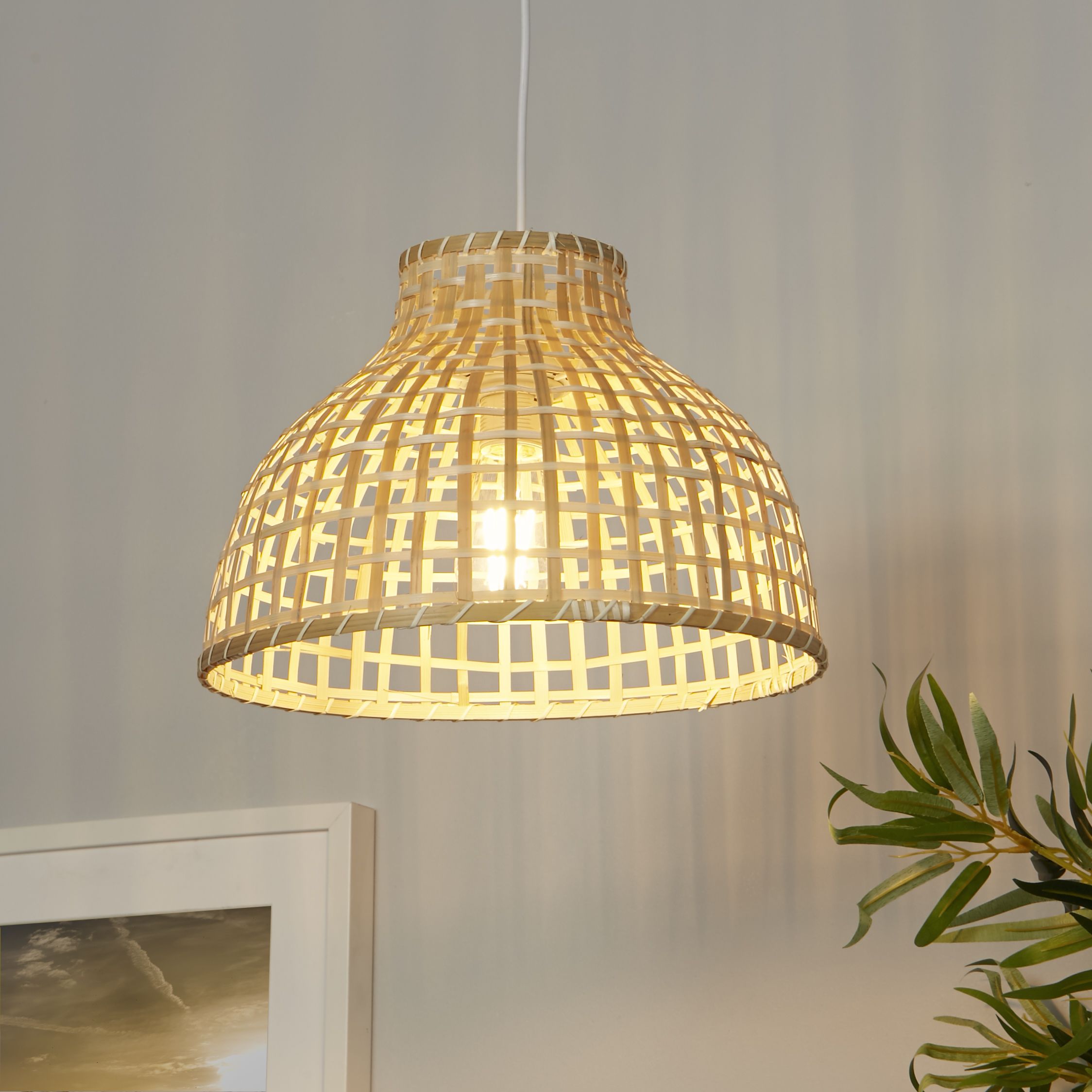 Bali Pendant in bamboo hanging in a styled room against a grey wall, beside a framed picture and green foliage