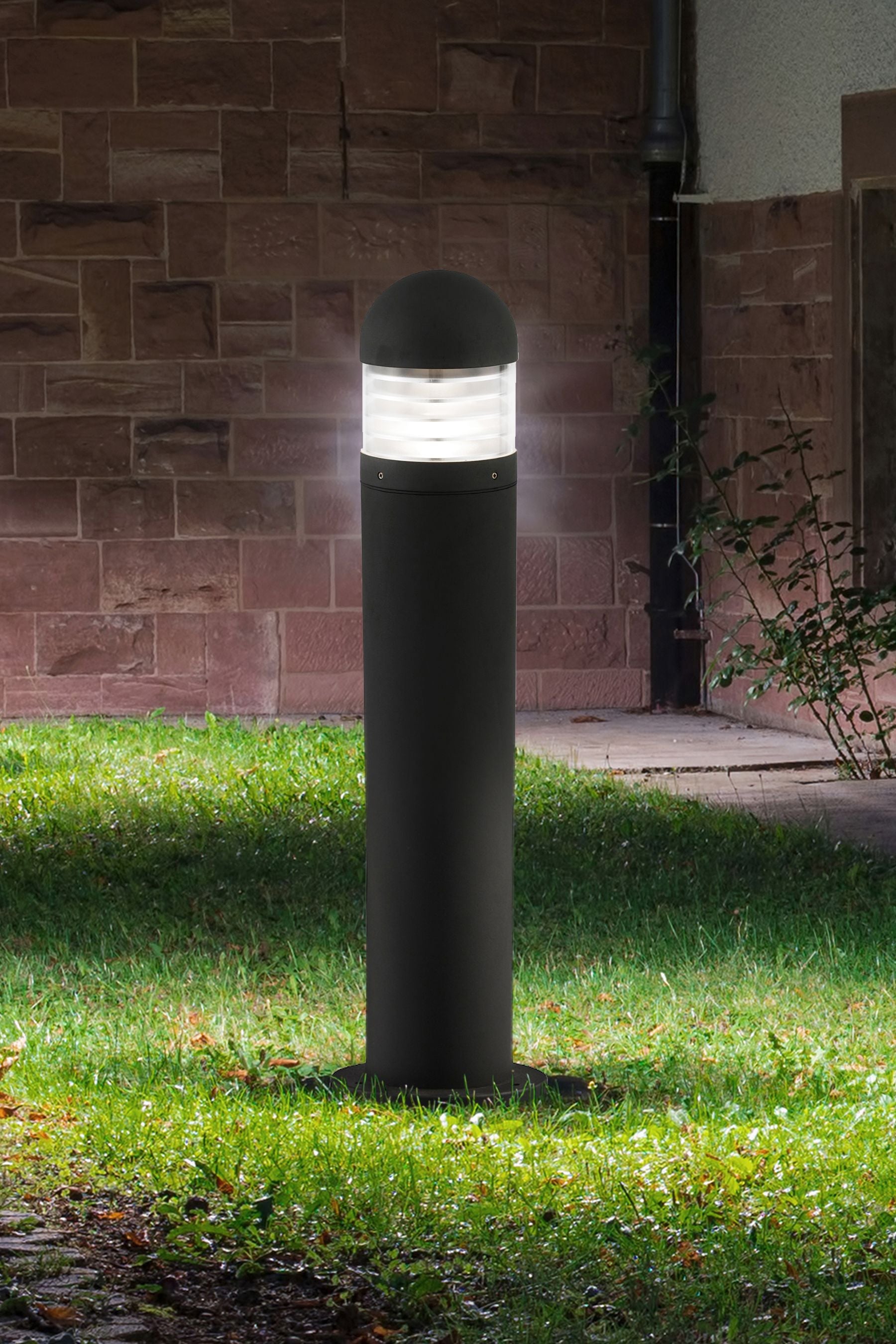 Bronx outdoor post light lit at night on a lawn beside a stone wall, illustrating the soft glow it casts around its base